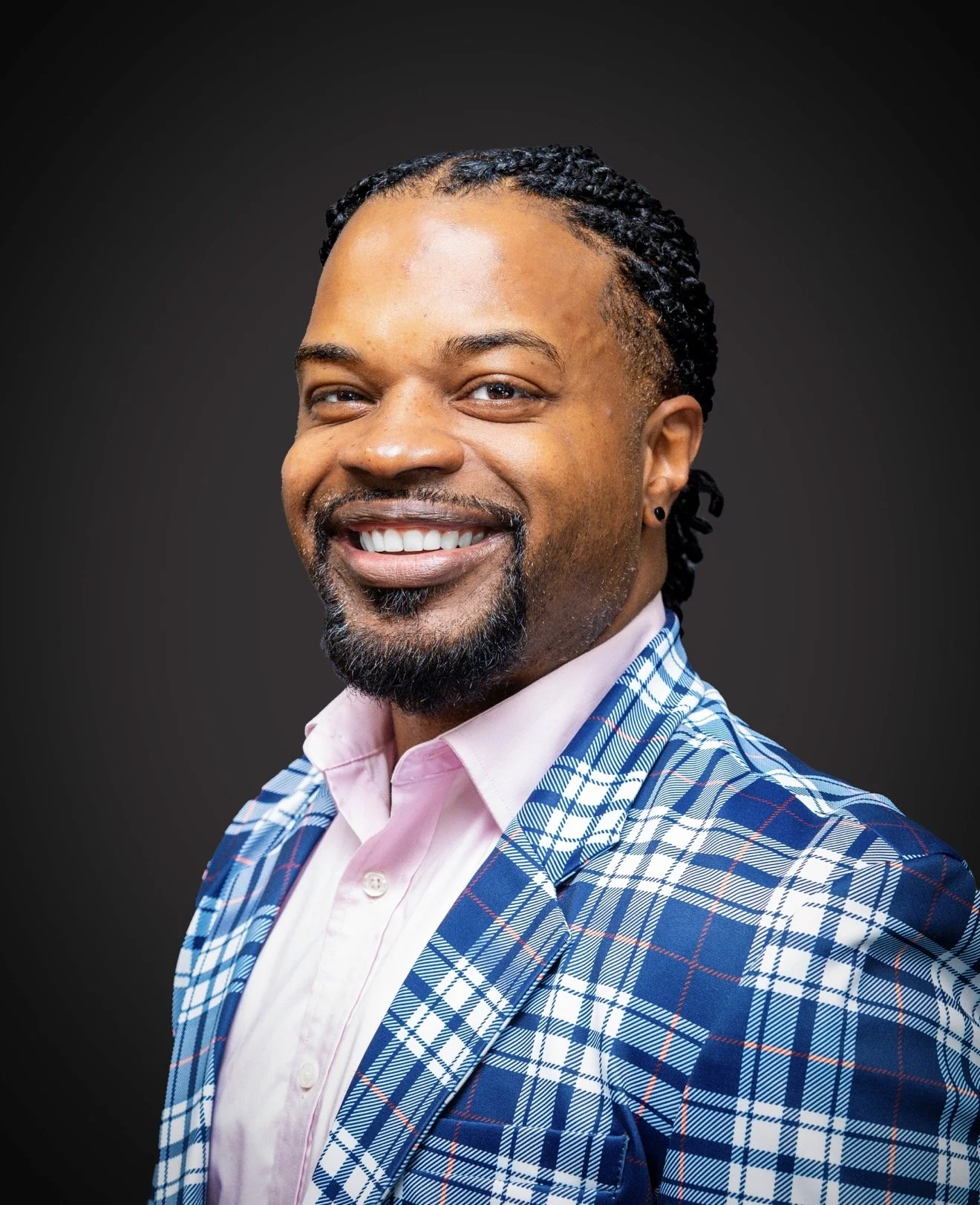 Smiling man with short dreadlocks, beard, wearing a pink shirt and a blue plaid blazer, against a dark background.