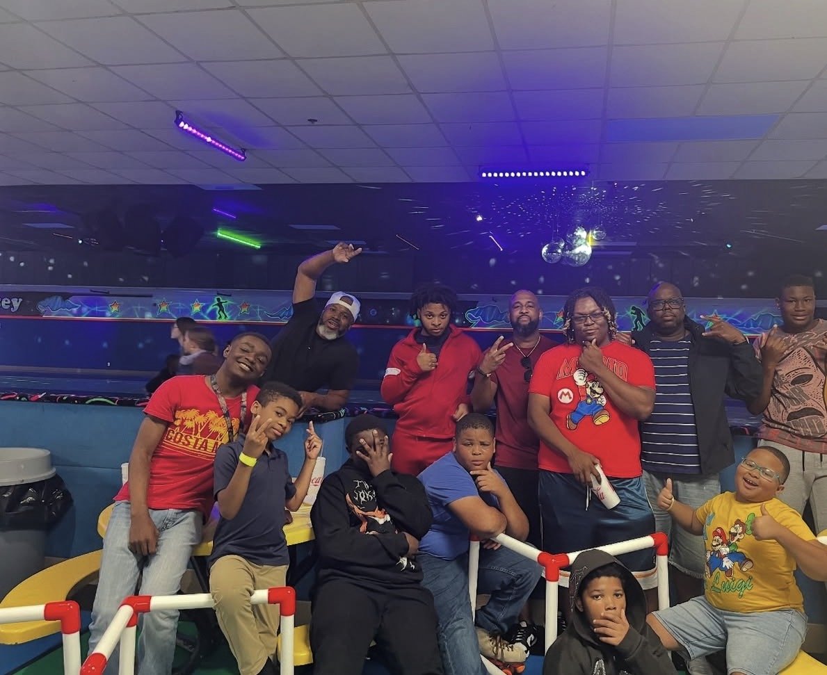 Group of kids and adults at a roller skating rink, with colorful lights and disco balls overhead, some making hand gestures, smiling, and posing for the camera.