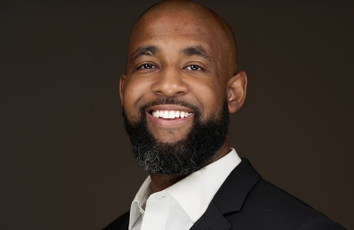 A smiling man with a bald head and a full beard wearing a dark suit and white shirt against a dark background.