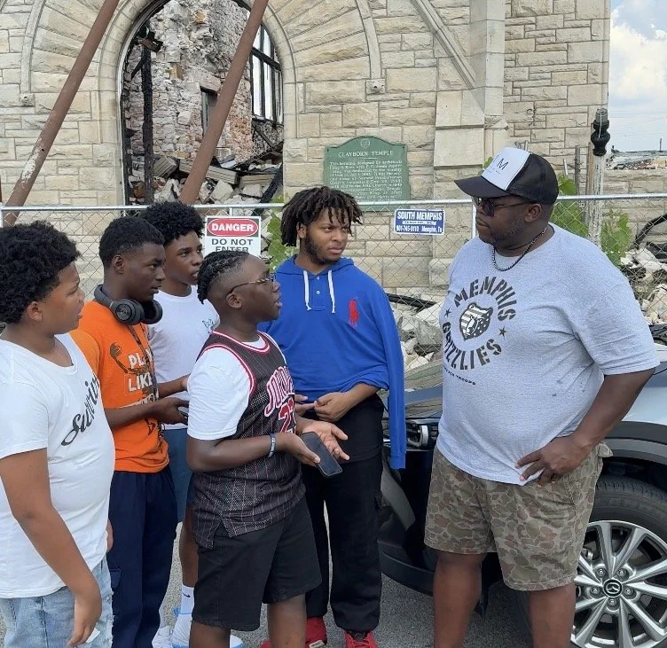 Group of young boys and an adult man standing outside near a damaged historic building with a chain-link fence, discussing or listening to each other.