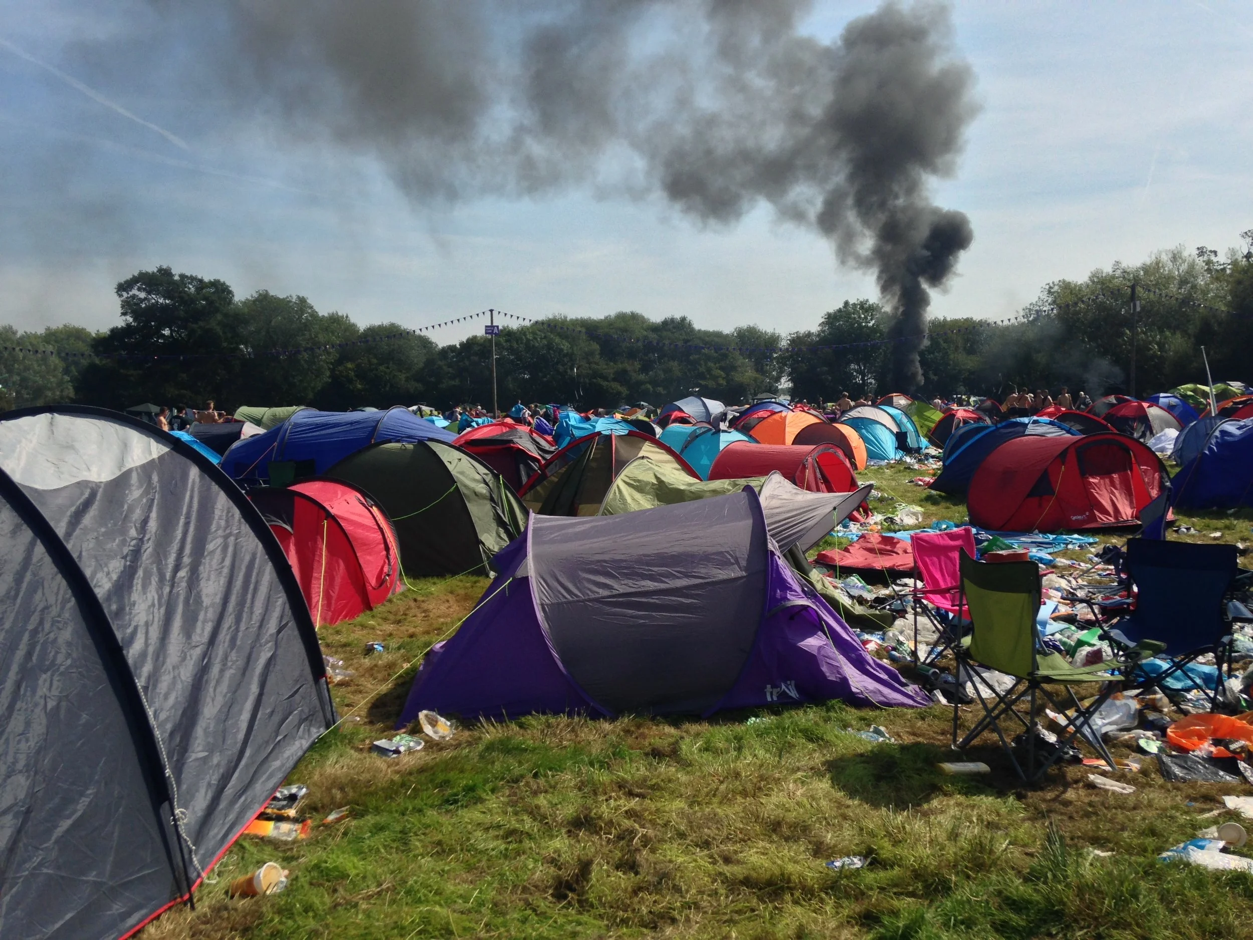 A camp area with numerous colorful tents set up on grass, some tents are overturned or disorganized. Smoke is rising in the background indicating a fire or explosion, with dark smoke billowing into the sky.