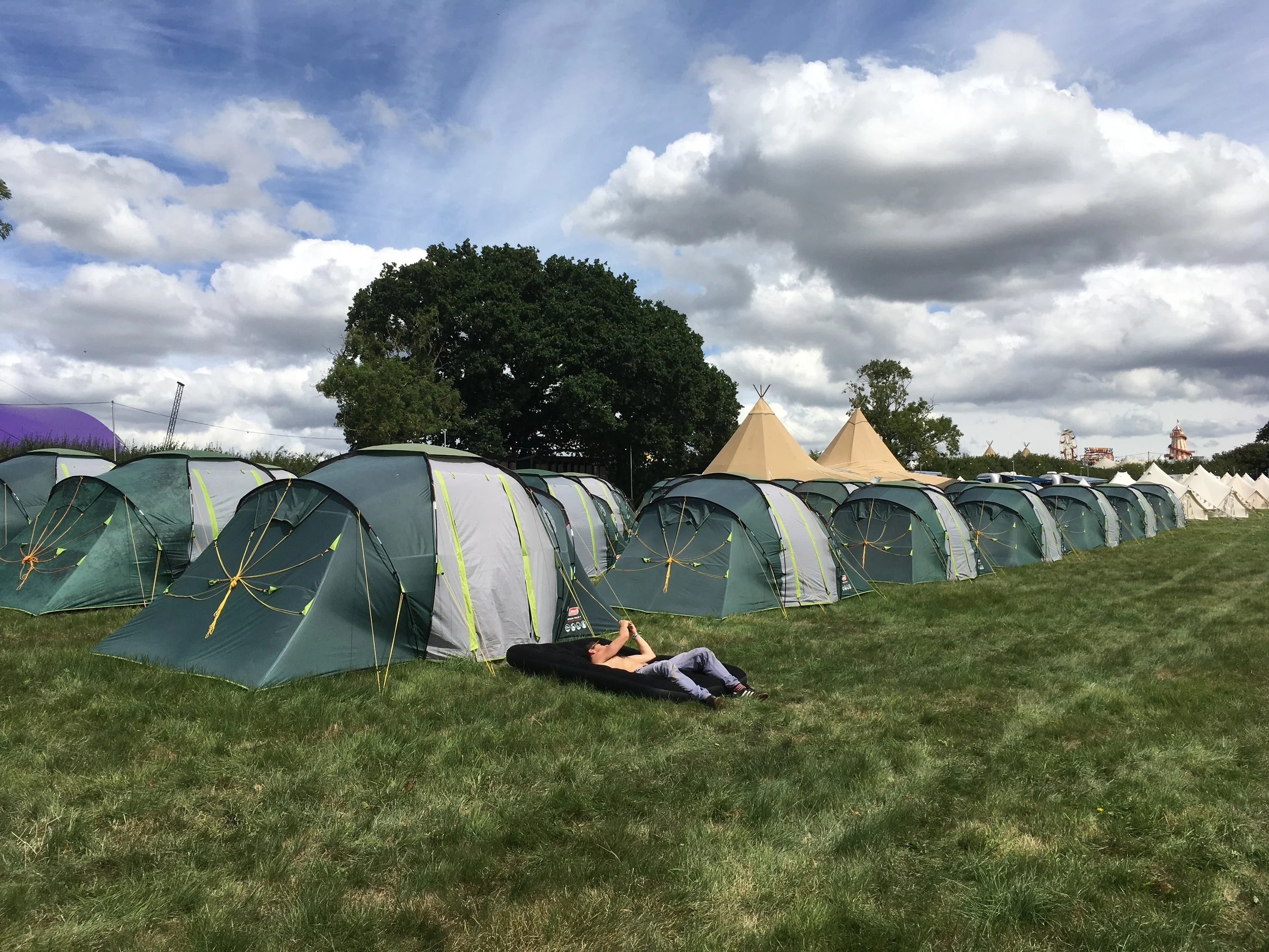 A person lying on a sleeping bag in front of a row of tents at an outdoor event, with a partly cloudy sky and trees in the background.