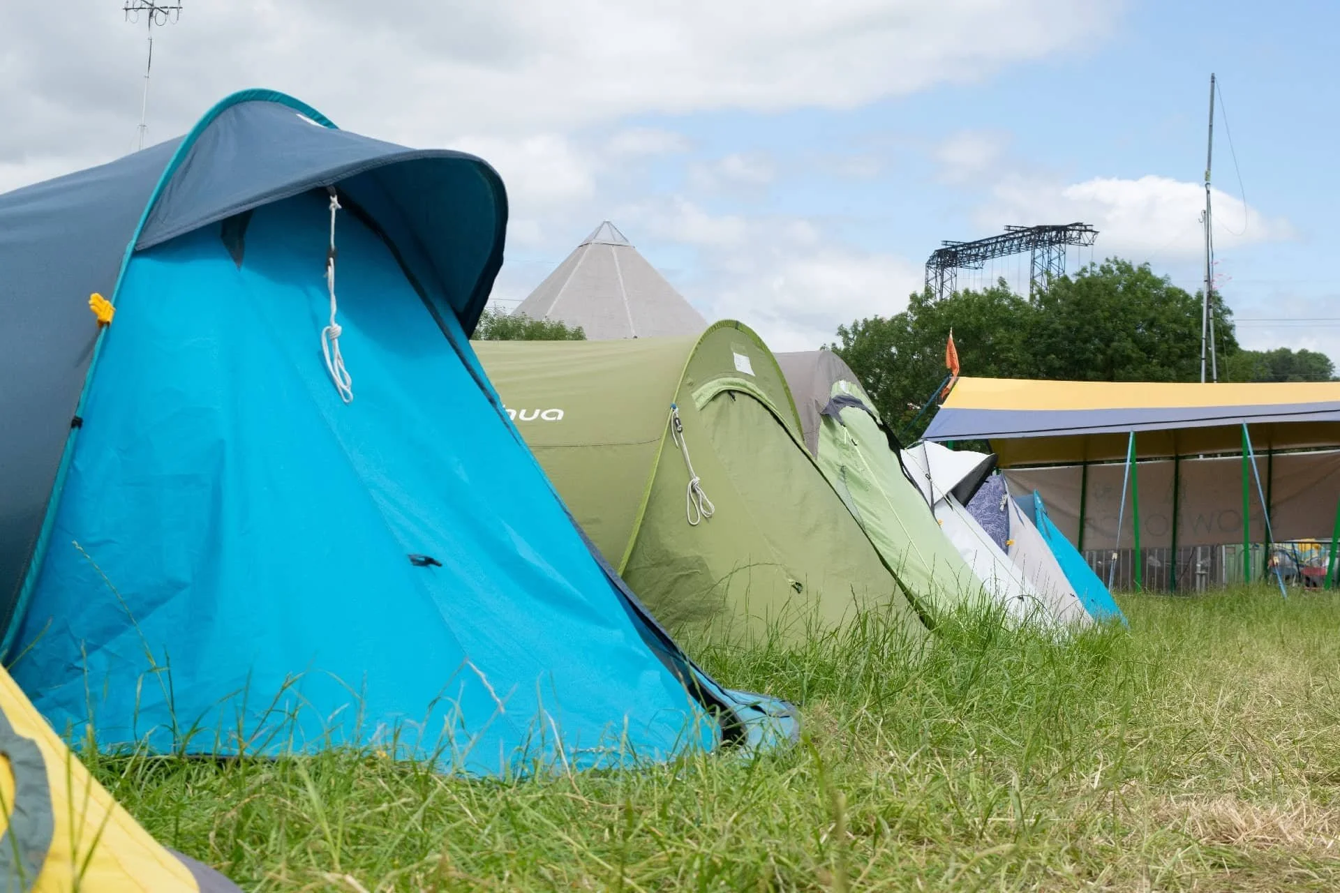 Multiple colorful tents set up on grass at an outdoor camping site with a pavilion and trees in the background.