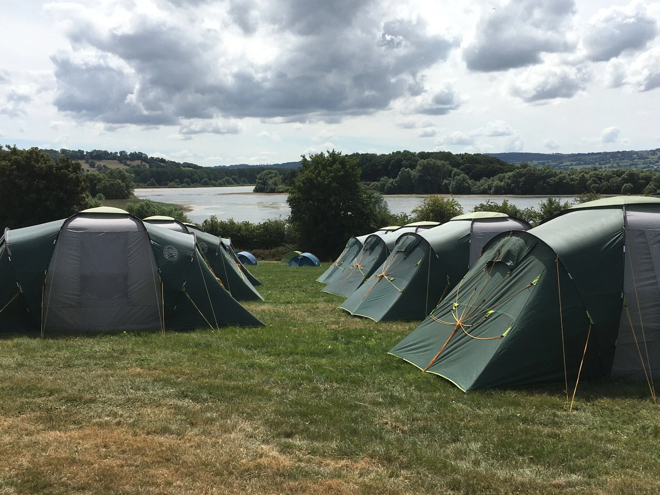Multiple green tents set up on a grassy area near water with trees and hills in the background under cloudy sky.