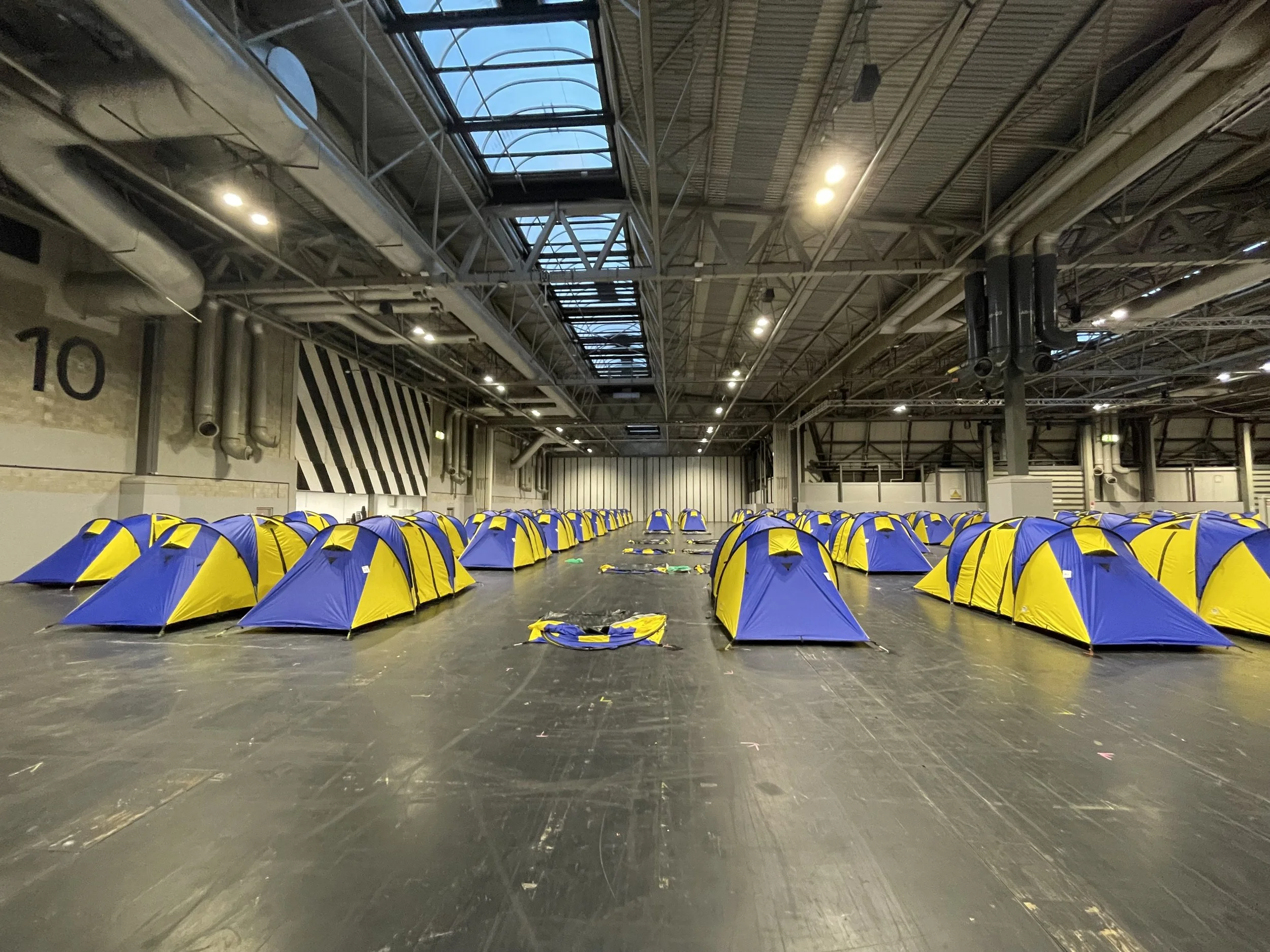 Rows of small blue and yellow tents set up inside a large industrial-style building with high ceilings and exposed pipes.