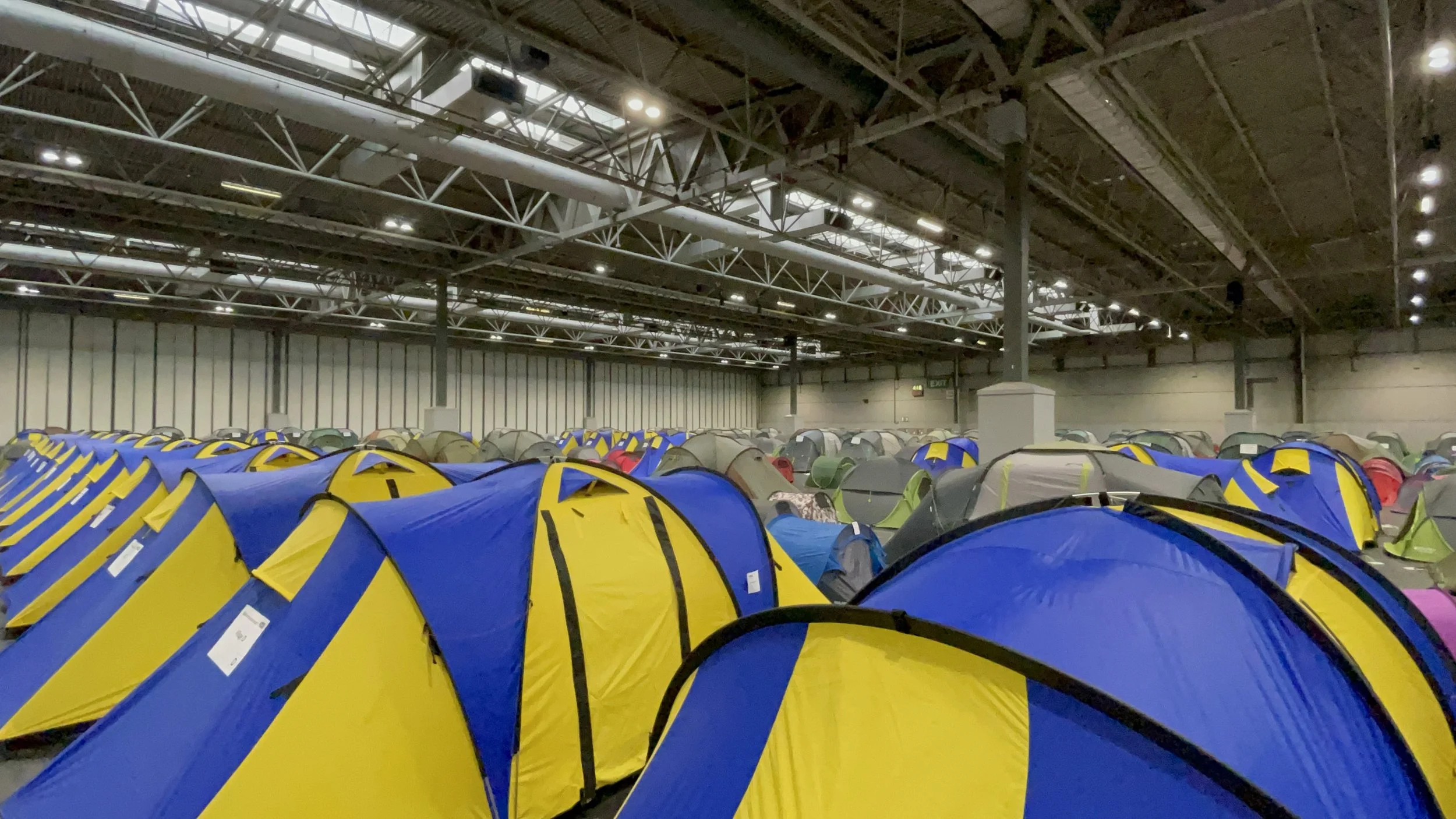 Indoor tent camping setup with numerous colorful tents, including blue and yellow, in a large warehouse or convention center with high ceiling and industrial lighting.