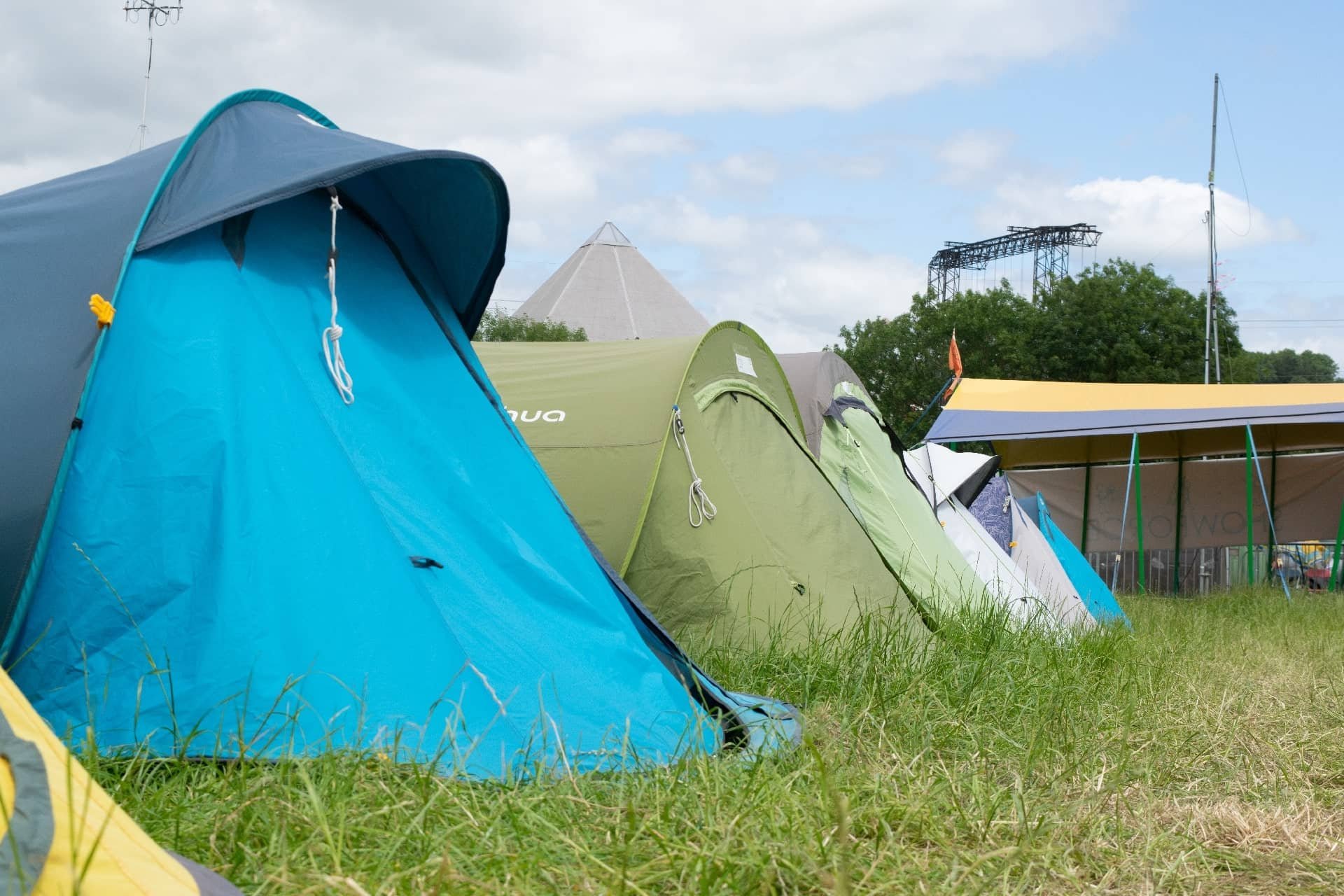 A row of colorful camping tents set up on grassy ground outdoors with a blue sky and some clouds, and trees in the background.