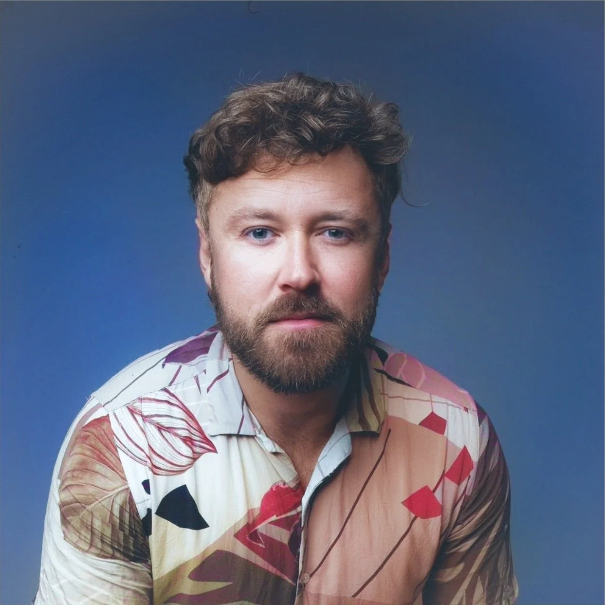 A man with curly brown hair, a beard, and blue eyes wearing a patterned short-sleeve shirt against a solid blue background.