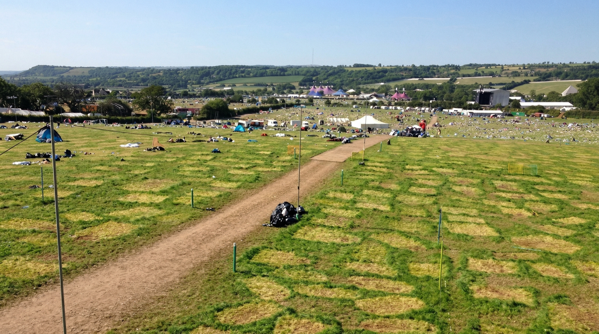 A large open grassy field with scattered tents and items, a dirt path running through it, and a stage in the distance with a crowd, surrounded by rolling hills and trees under a blue sky.