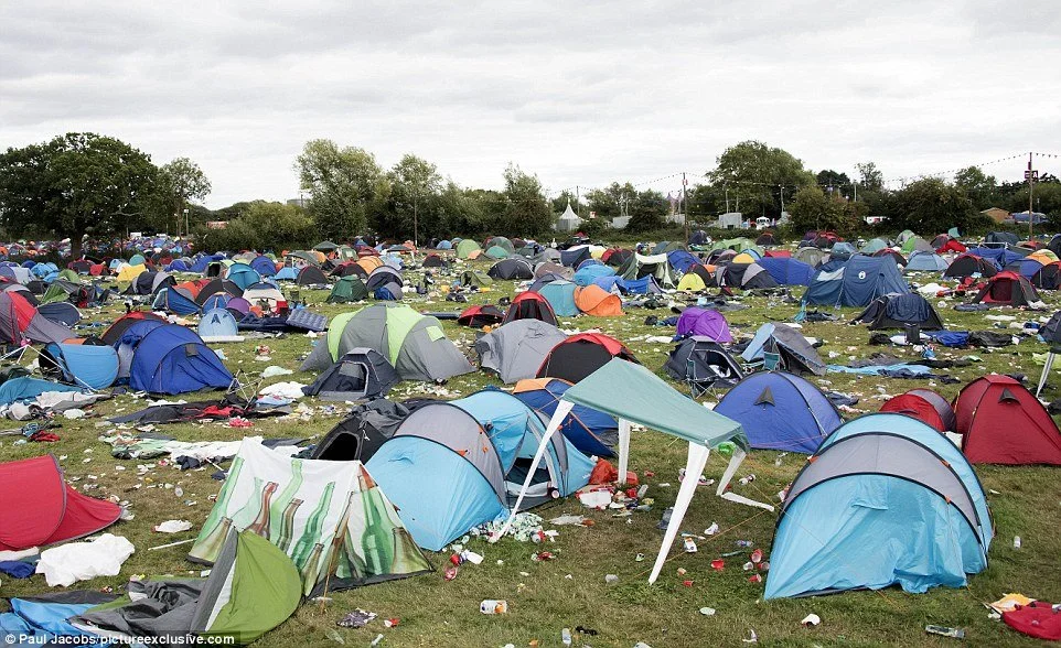 A large encampment with numerous tents scattered across a grassy field, with trash and debris around. Overcast sky and trees in the background.