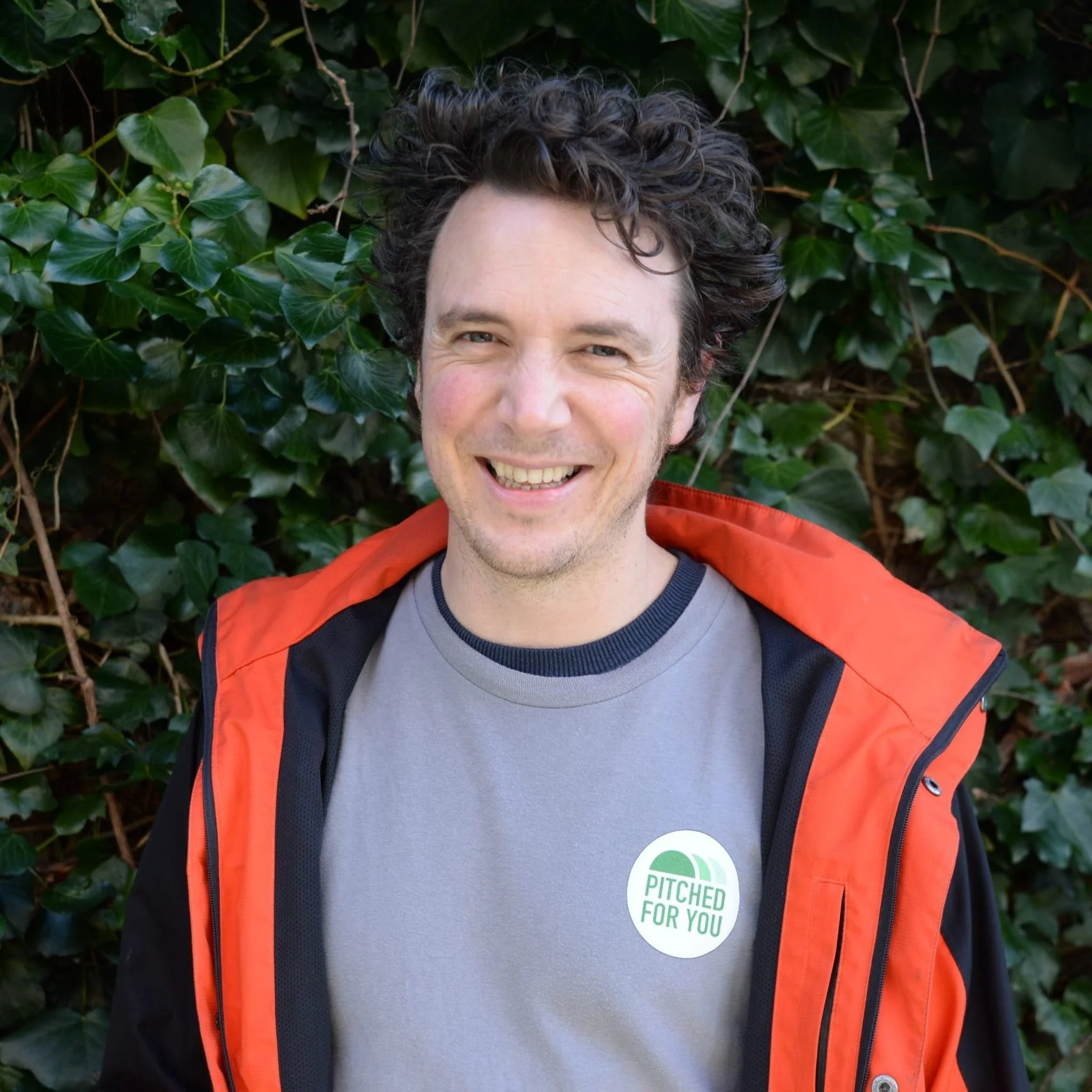 A smiling man with dark, curly hair standing in front of green foliage, wearing a gray shirt with a green and white sticker that says 'Pitched For You' and an orange jacket.