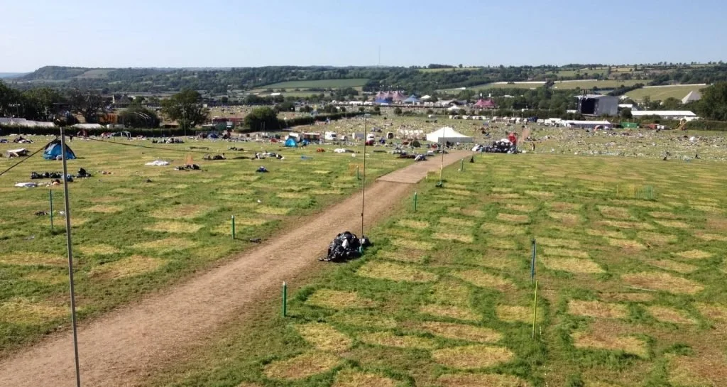 Crowd lying on grass fields at an outdoor event with a stage and tents in the background.