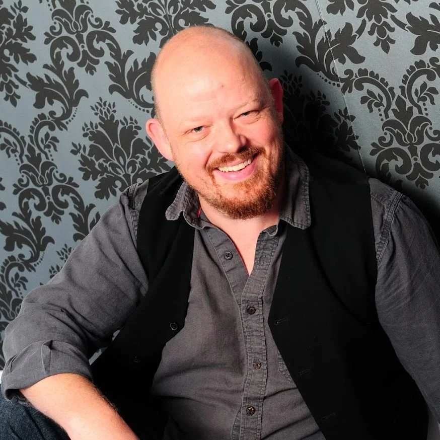 A smiling man with a beard and short hair, wearing a gray shirt and black vest, sitting against a gray and black floral-patterned wall.