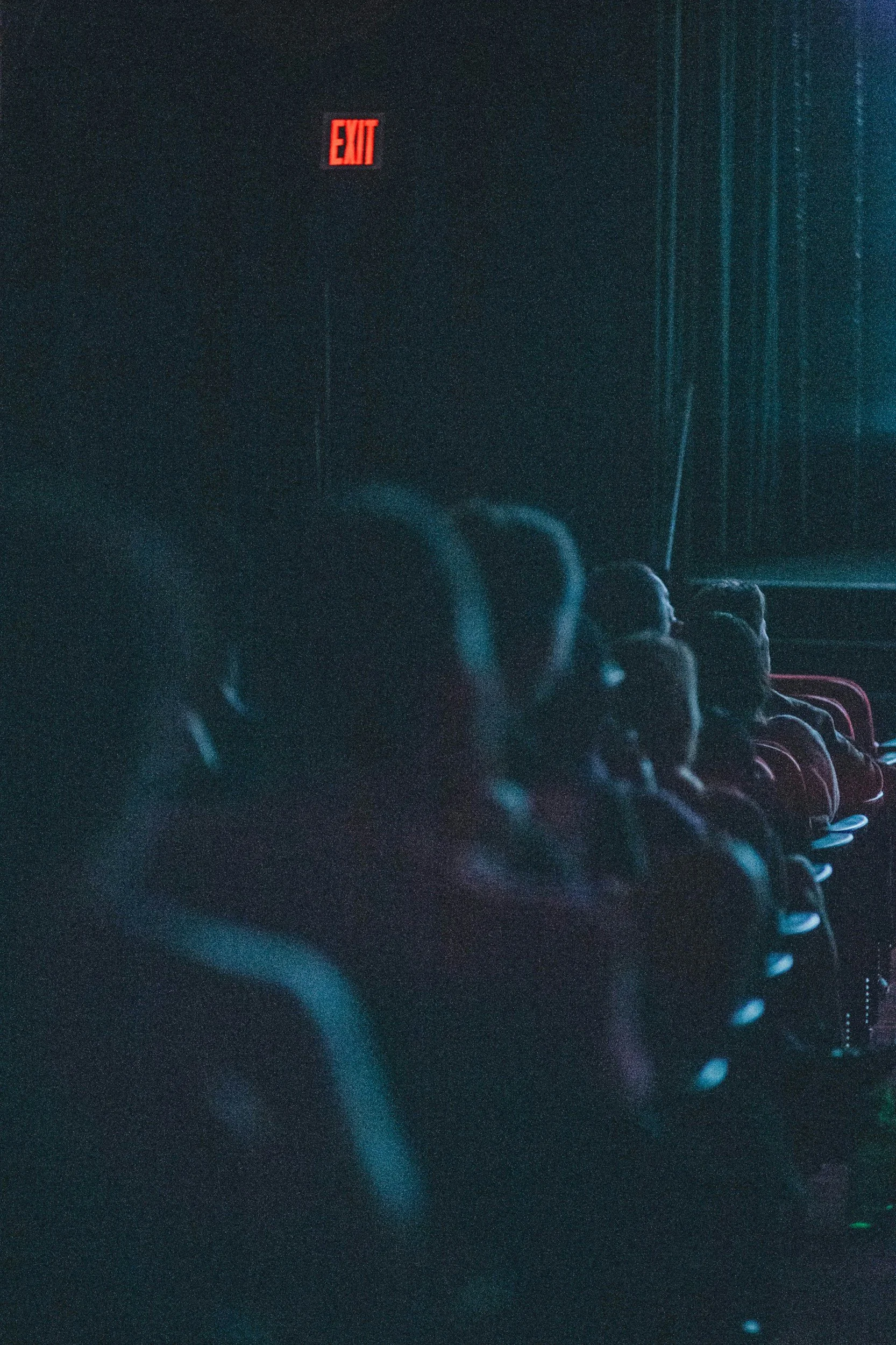A dark cinema auditorium filled with people seated in rows, with a lit red exit sign visible on the wall - The Power of Story - Discover Narrative Coaching.
