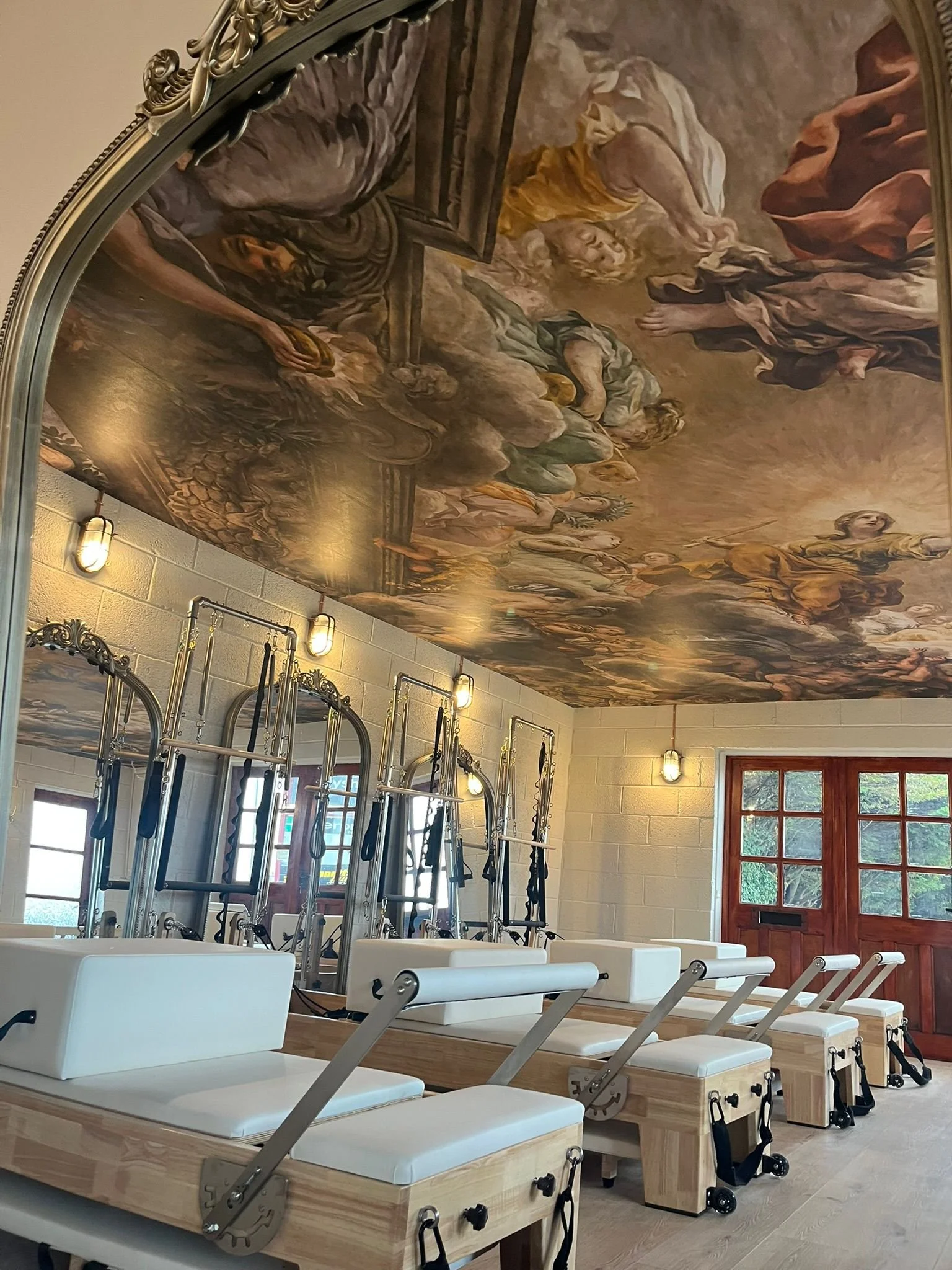 Pilates reformer equipment in a room with a painted ceiling depicting a classical scene, illuminated by wall-mounted lights and large windows.