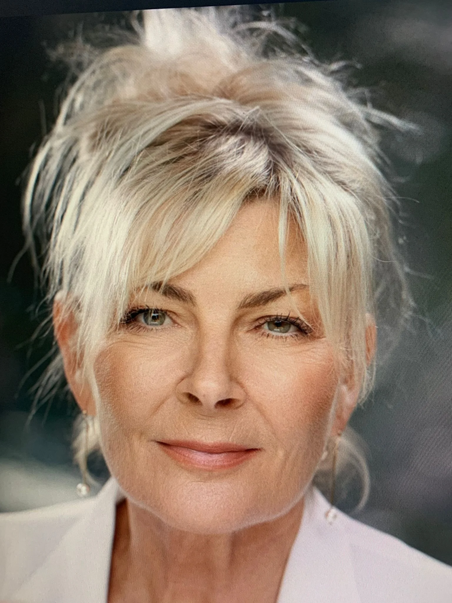 A close-up portrait of an older woman with short, styled, blonde hair and light-colored eyes, smiling softly, wearing light makeup and earrings.