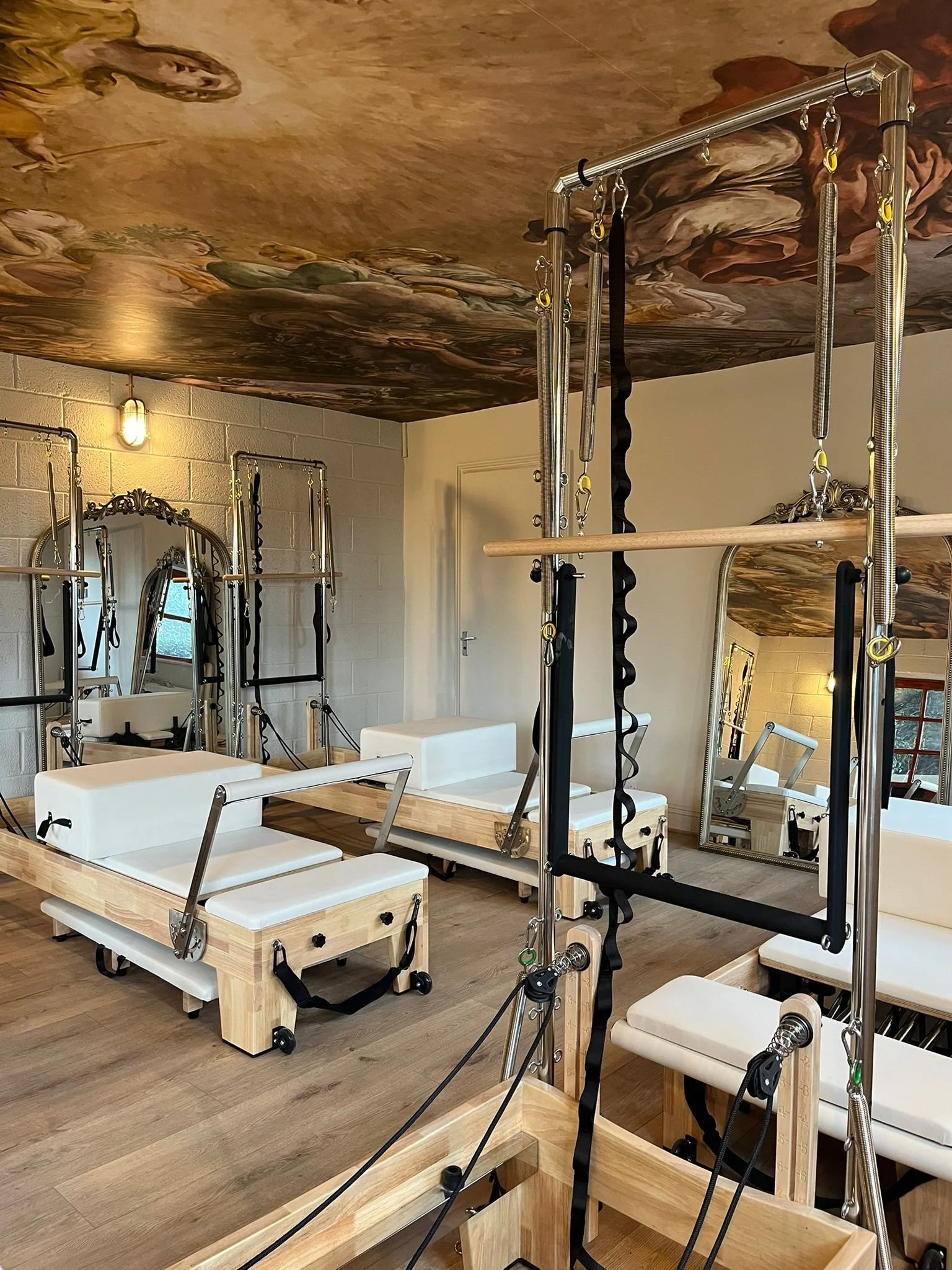 Pilates reformer equipment with wooden frames, padded surfaces, and metal bars in a studio with mirrors and wood floors.