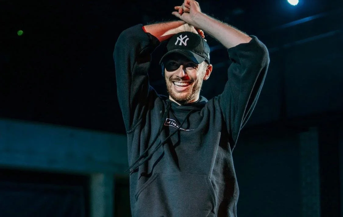 A man wearing a black baseball cap with a white logo, sunglasses, and a dark hoodie, smiling and raising his hands above his head in a dimly lit indoor setting.