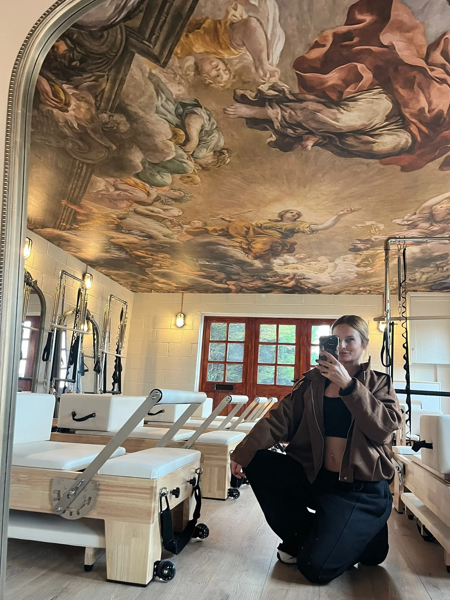 A woman is taking a mirror selfie in a Pilates or physical therapy studio with wooden equipment and a ceiling painted with a classical fresco scene.