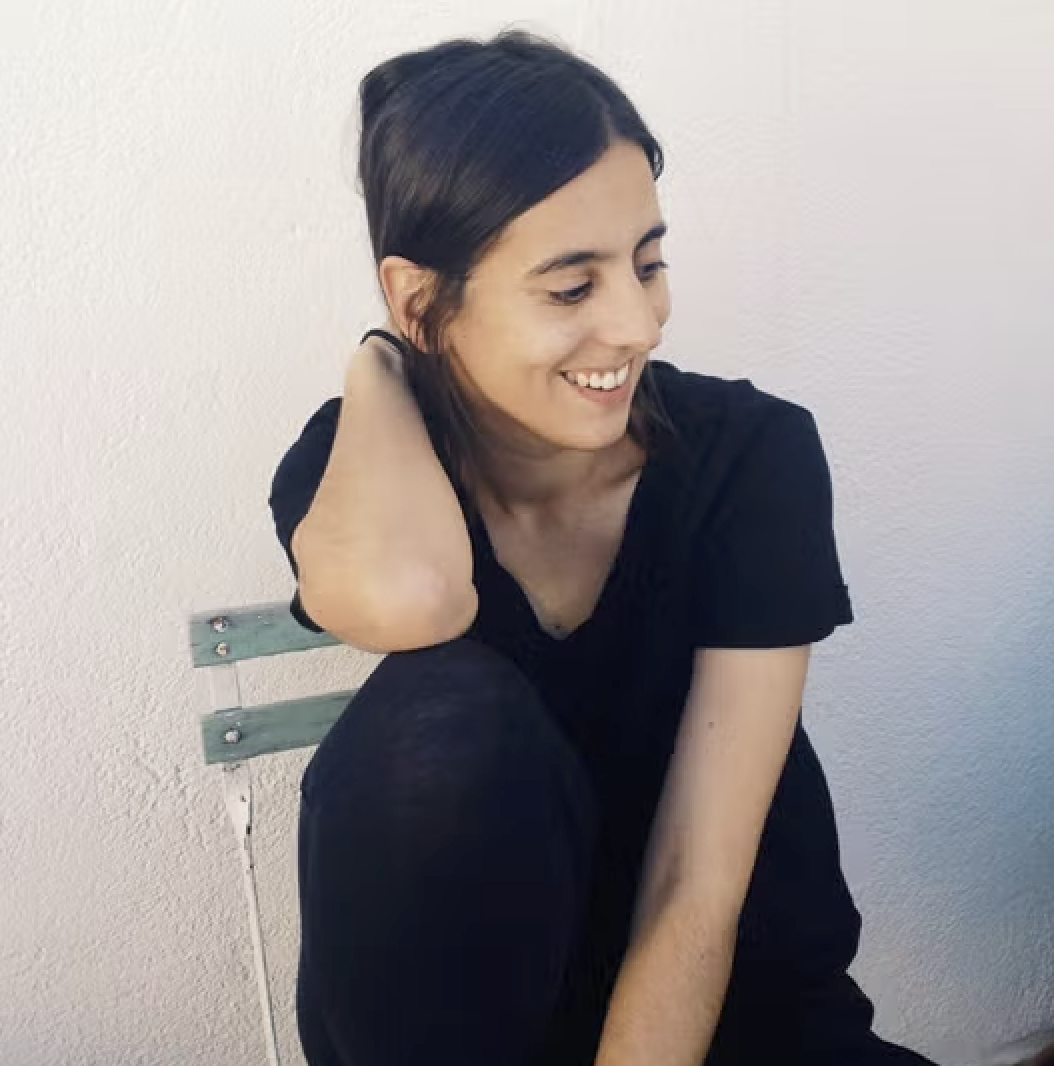 Mujer joven sonriendo, sentada en una banca de pared blanca.