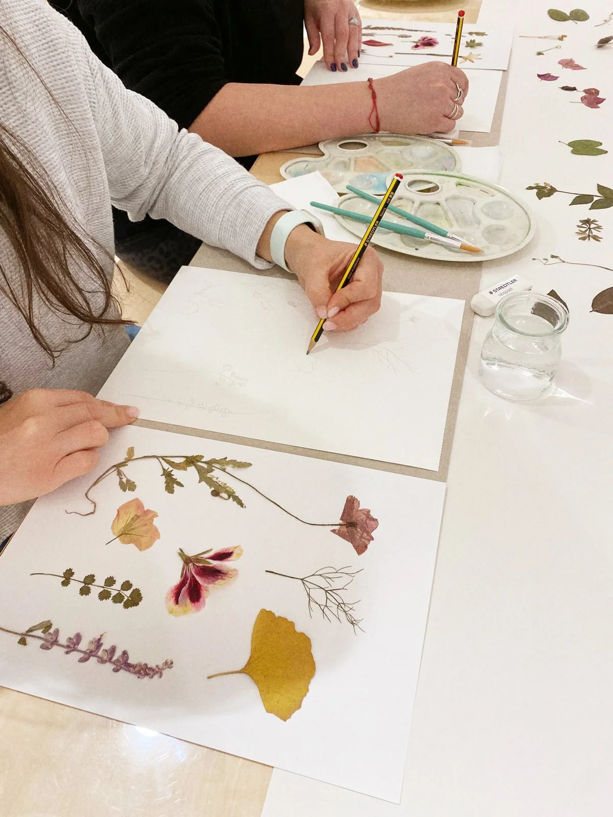 Personas creando arte con hojas secas y flores prensadas en papel en una mesa de pintura.