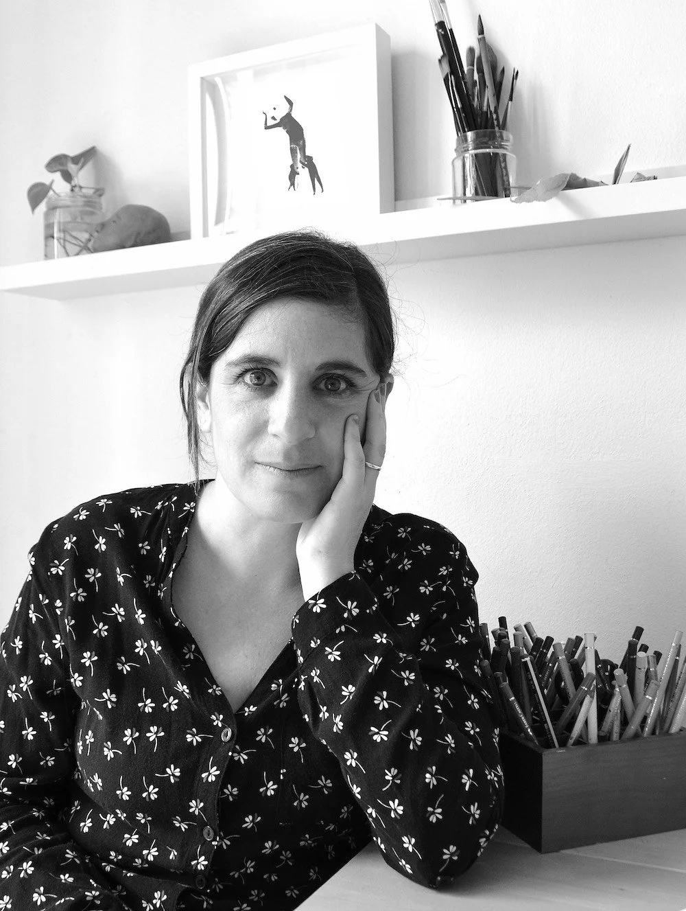 Retrato de una mujer con cabello oscuro y piel clara, usando una blusa negra con estampado de pequeñas flores blancas, sentado en un escritorio en una oficina o estudio, con estanterías blancas y objetos decorativos en el fondo.