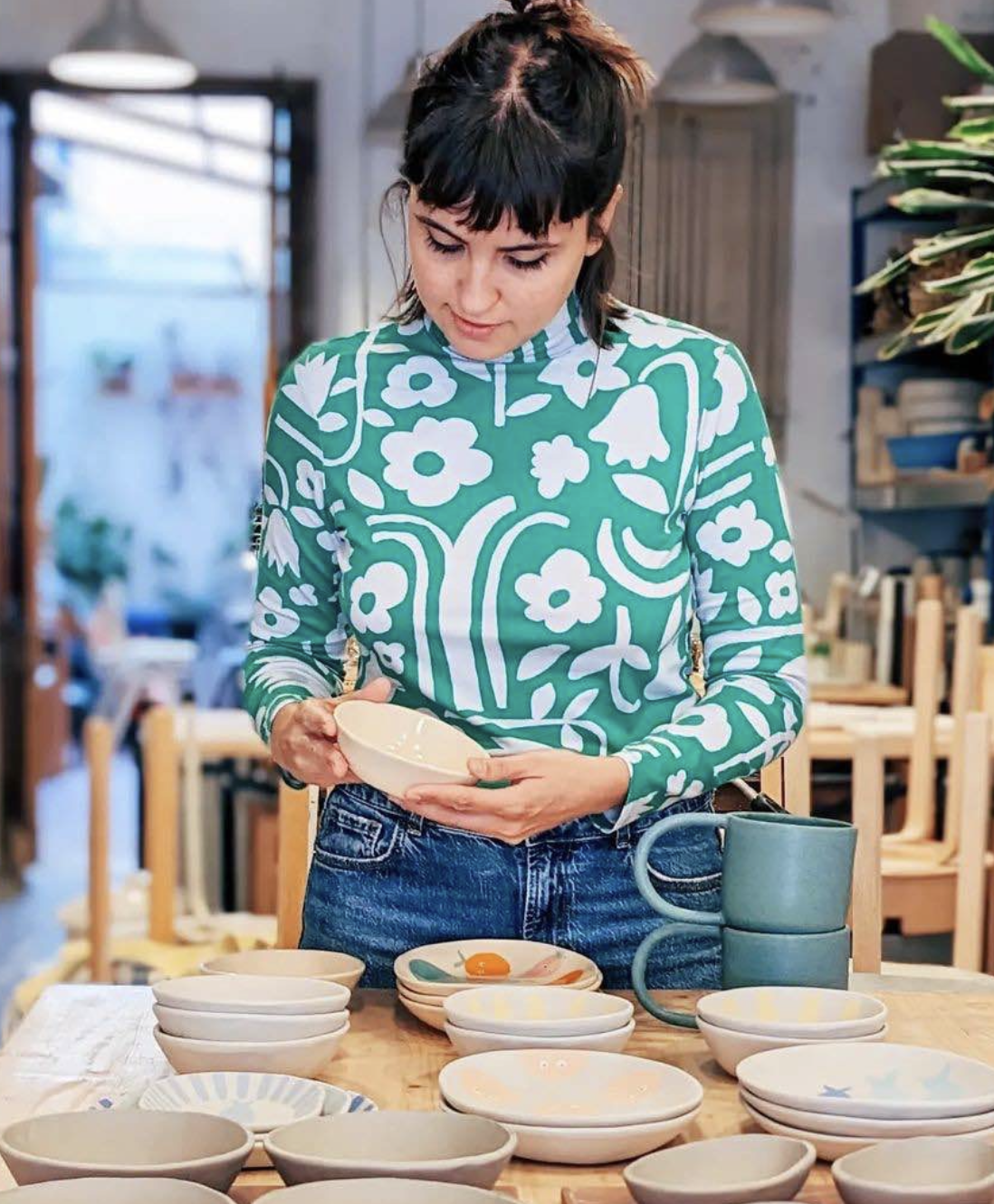 Mujer sosteniendo un plato pequeño en una mesa de cerámica, rodeada de otros tazones y platos. Ella viste una blusa de manga larga con estampado de flores en tonos verdes y blancos, y jeans azules. El ambiente parece ser una tienda o taller de cerámica.