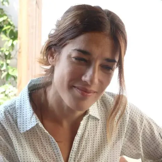 Mujer con expresión de incomodidad o duda, con cabello castaño y blusa blanca con puntos, en un ambiente con luz natural y fondo de madera y plantas.