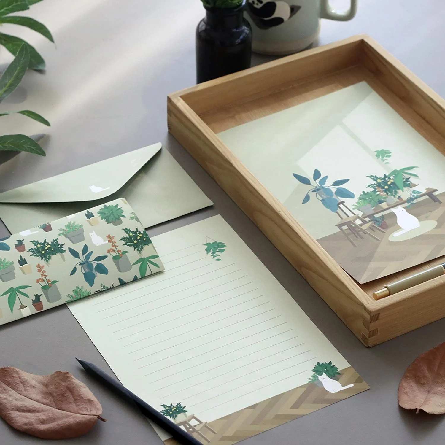 Estación de escritorio con papelería decorada con temas de plantas, una pluma y un reconocimiento de escritorio con ilustraciones de plantas y una gata blanca, sobre una superficie gris, con hojas secas y una planta en el fondo.
