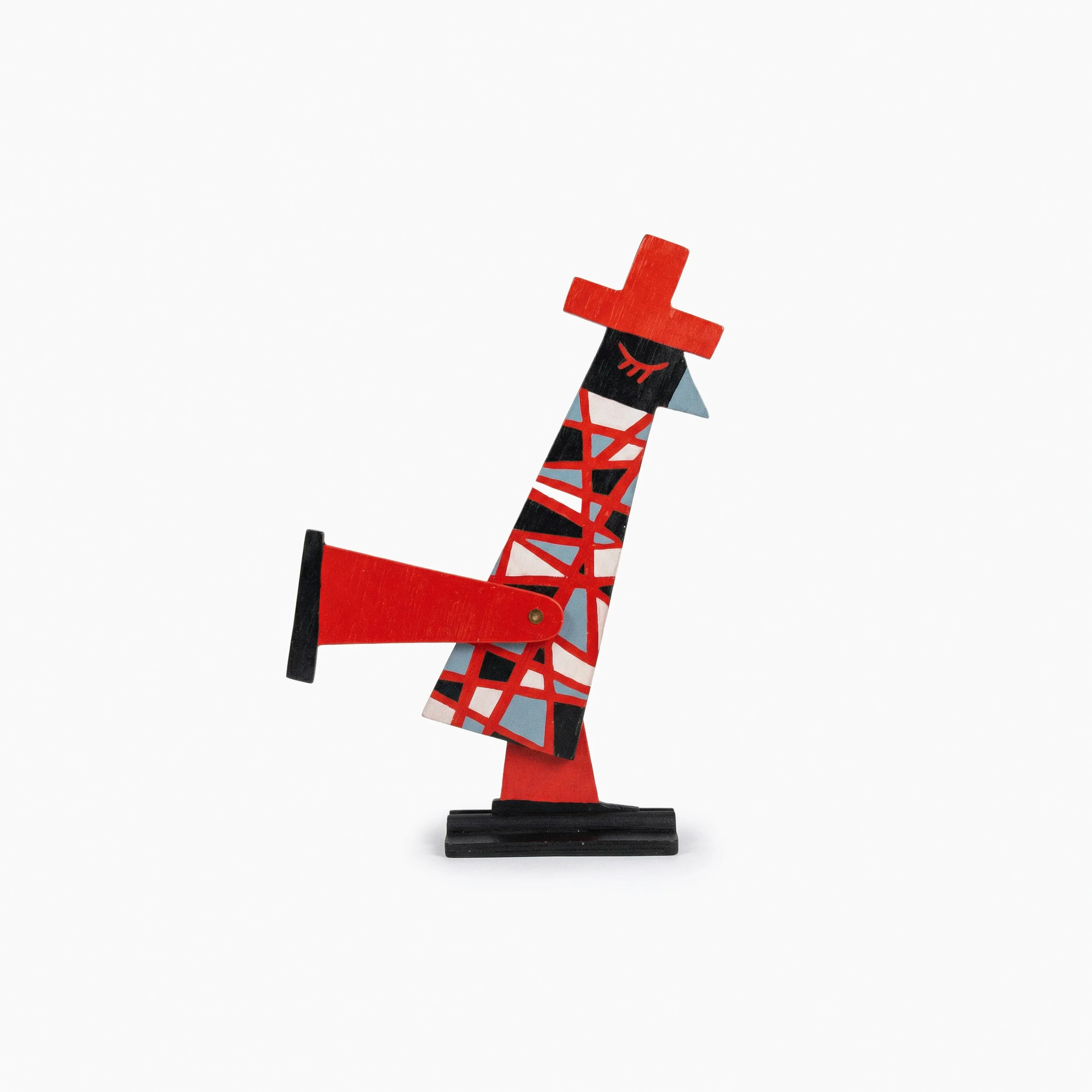 Figura de madera con forma de jirafa decorada con patrones de colores rojo, negro, gris y blanco, con una cabeza en forma de cruz roja y ojos cerrados, sobre un fondo blanco y simple.