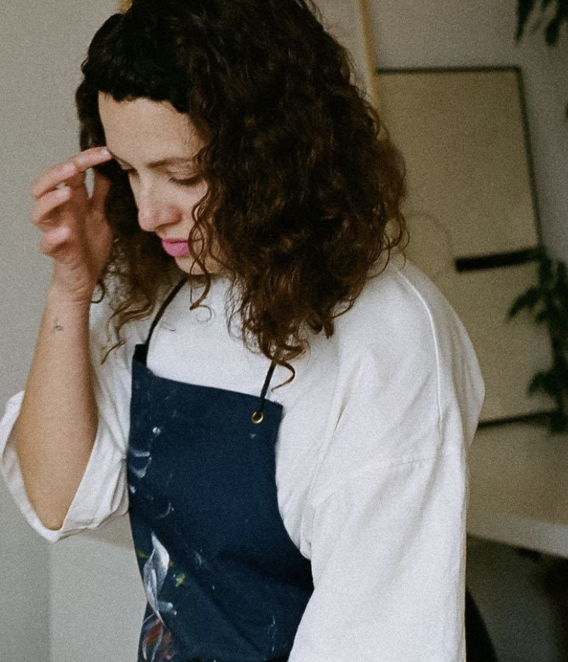 Mujer con cabello rizado, vestida con camiseta blanca y delantal azul marino, tocándose la cabeza en un ambiente interior.