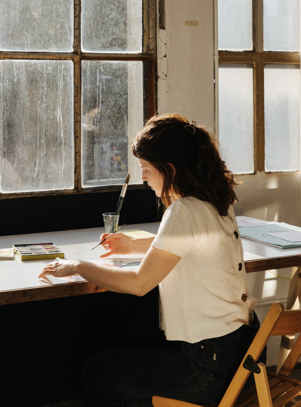 Una mujer con cabello castaño oscuro pintando en un estudio, con luz natural entrando por ventanas viejas y conectando con un ambiente artístico.