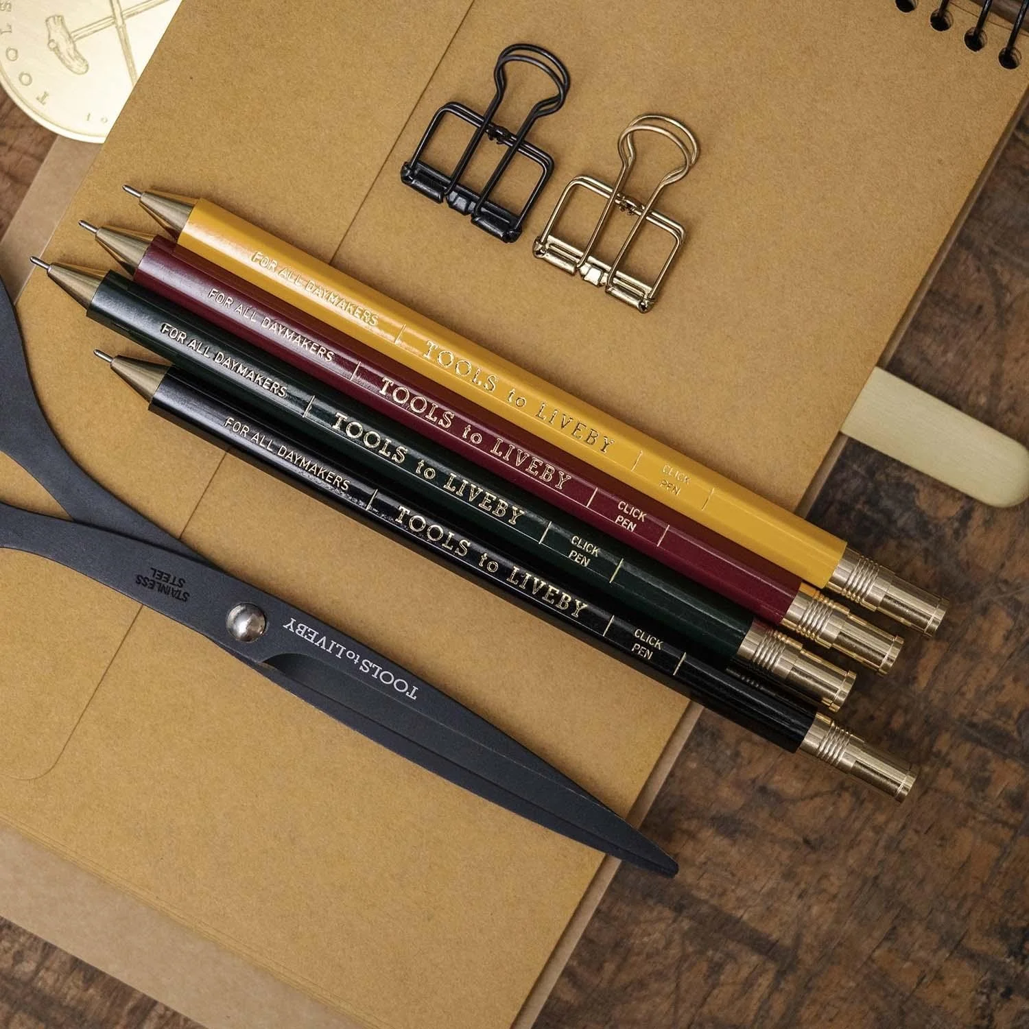 Cuatro bolígrafos de diferentes colores (amarillo, burdeos, verde y negro), dos sujetapapeles en tonos dorados y negros, una cinta de medición gris con la marca 'TOOLS to LIVEBY', y un cuaderno de notas de tapa de cartón en color marrón claro.