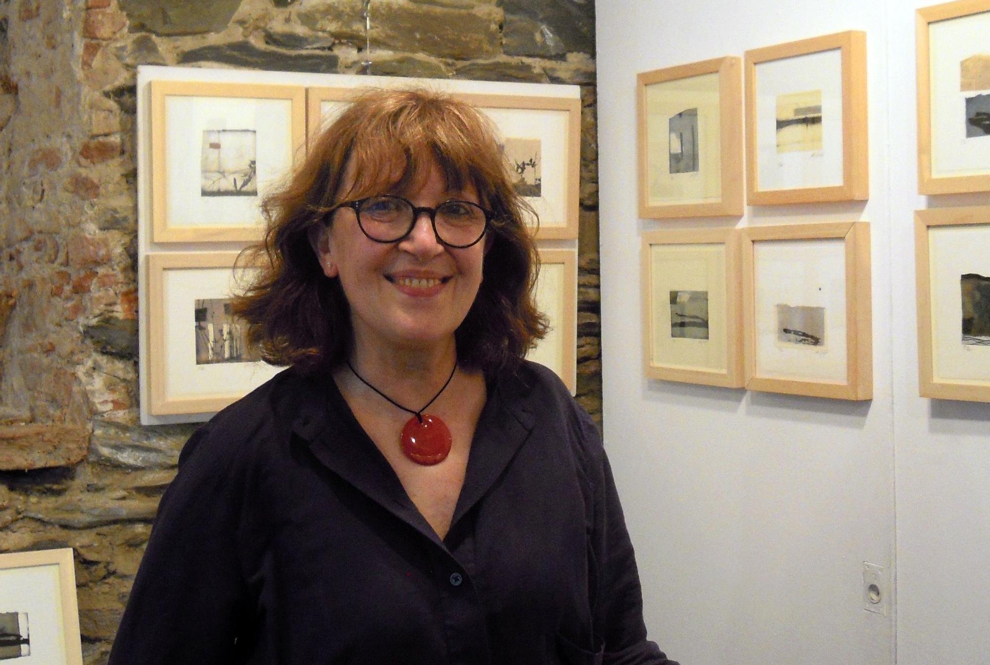 Una mujer con gafas y collar rojo, sonriendo, en una galería de arte con paredes de piedra y cuadros enmarcados en fondo claro.