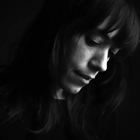 Retrato en blanco y negro de una mujer con cabello largo y oscuro, mirada hacia abajo, en un ambiente oscuro.