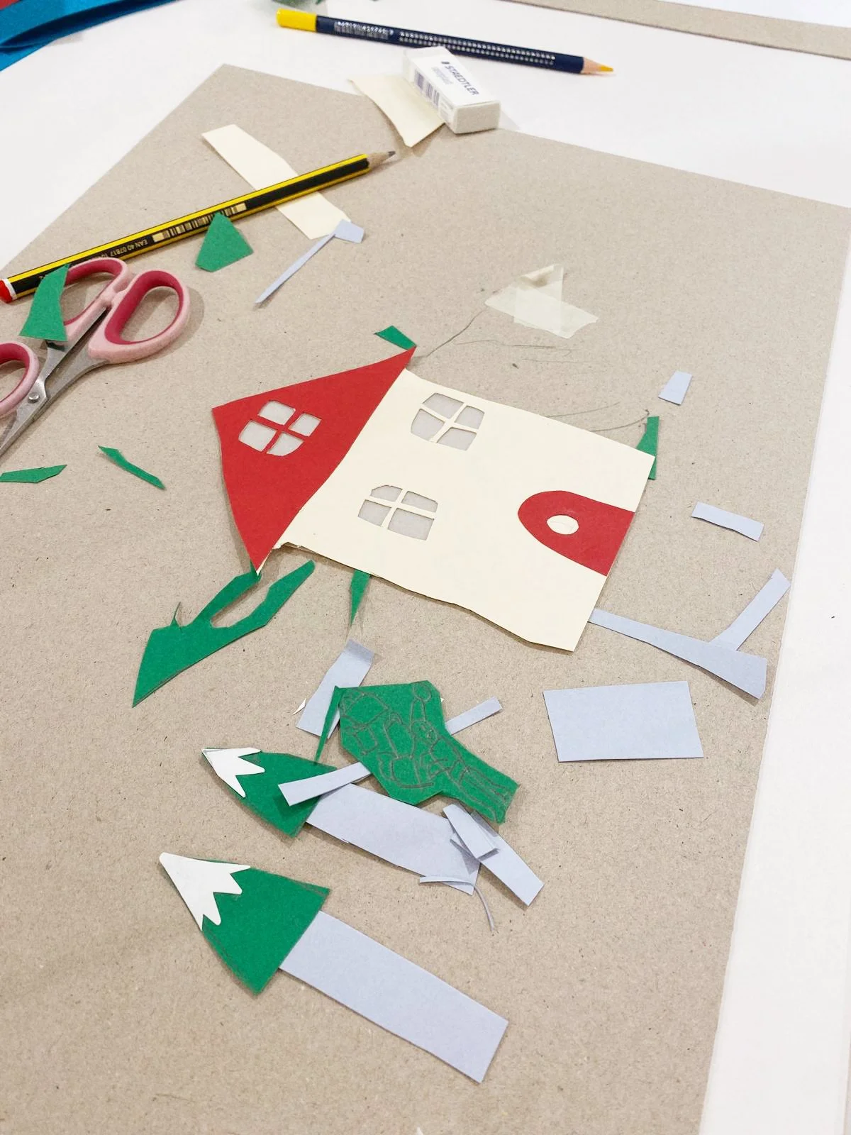 Material de manualidades con recortes de papel, tijeras, cinta, y un cascanueces de papel que forma una casita de Navidad en proceso de construcción.