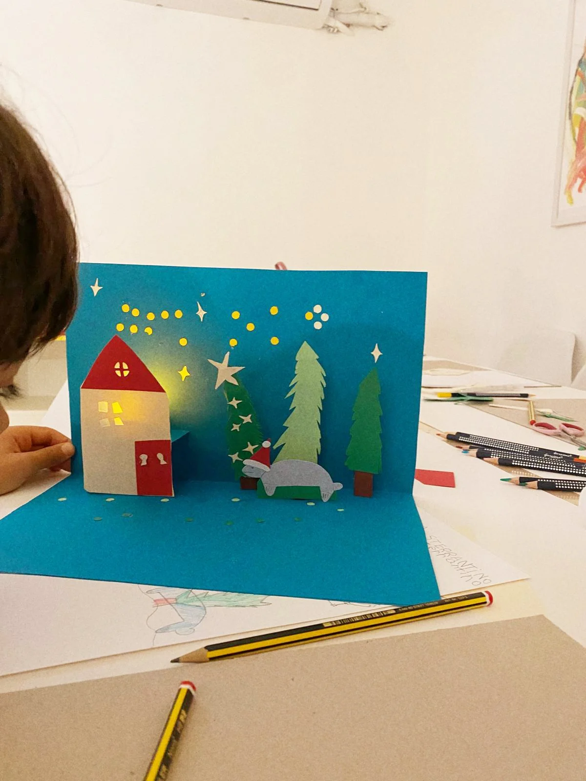 Tarjeta de Navidad con árboles, una casa y un reno con gorro, en un fondo azul, decorada con estrellas y puntos de colores