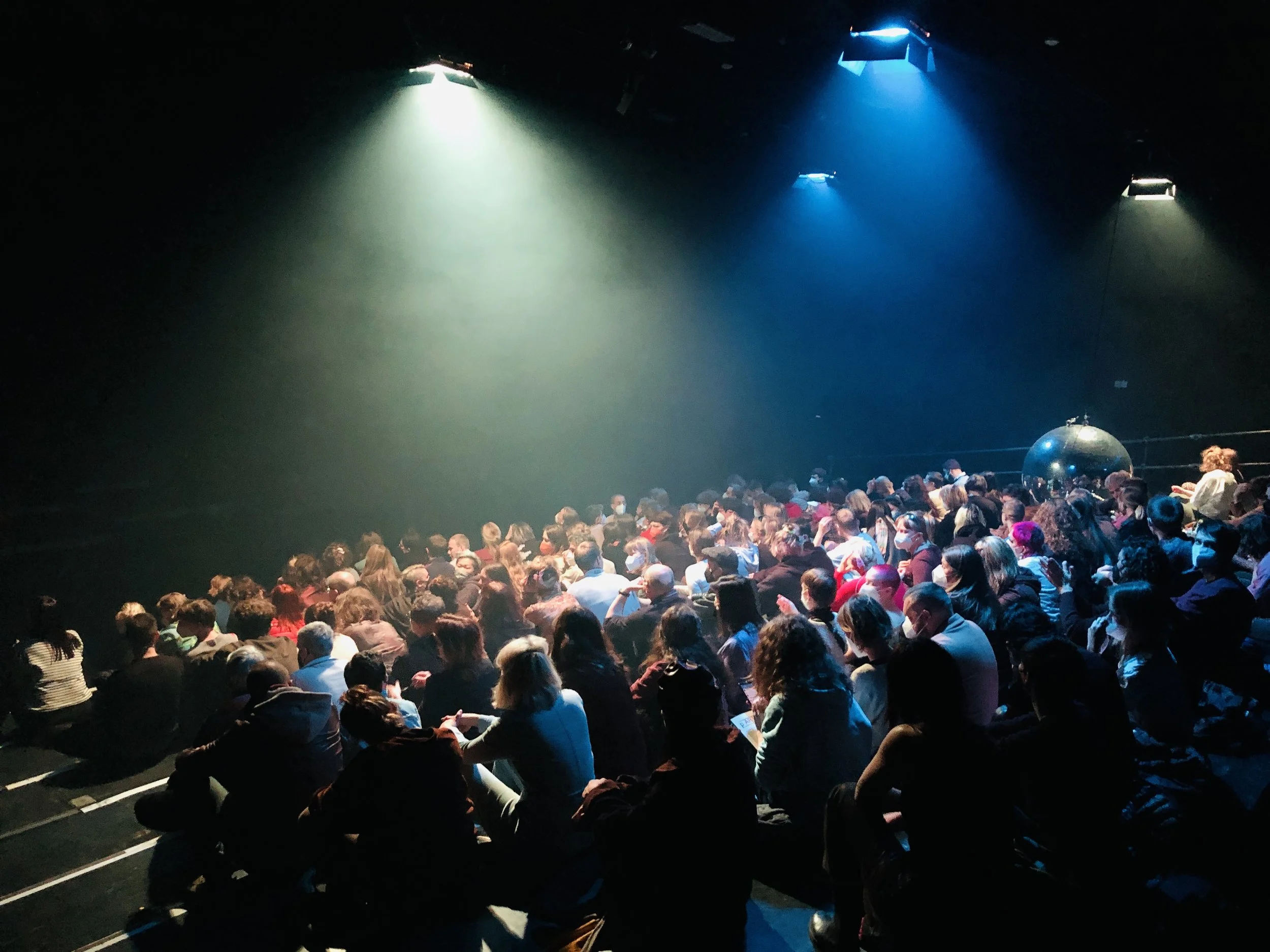 A large audience seated in a dark theater or auditorium, illuminated by a few colored spotlights overhead.