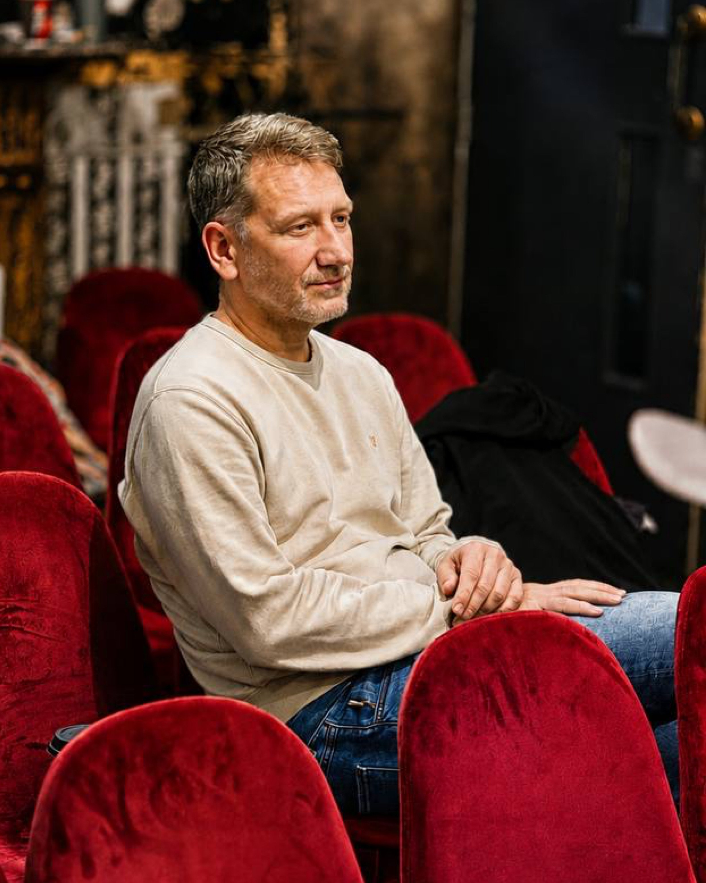 A middle-aged man with gray hair and a beard, wearing a beige sweatshirt, sitting in a red velvet seat in a theater or cinema, with others seated nearby and a dark, ornate background.