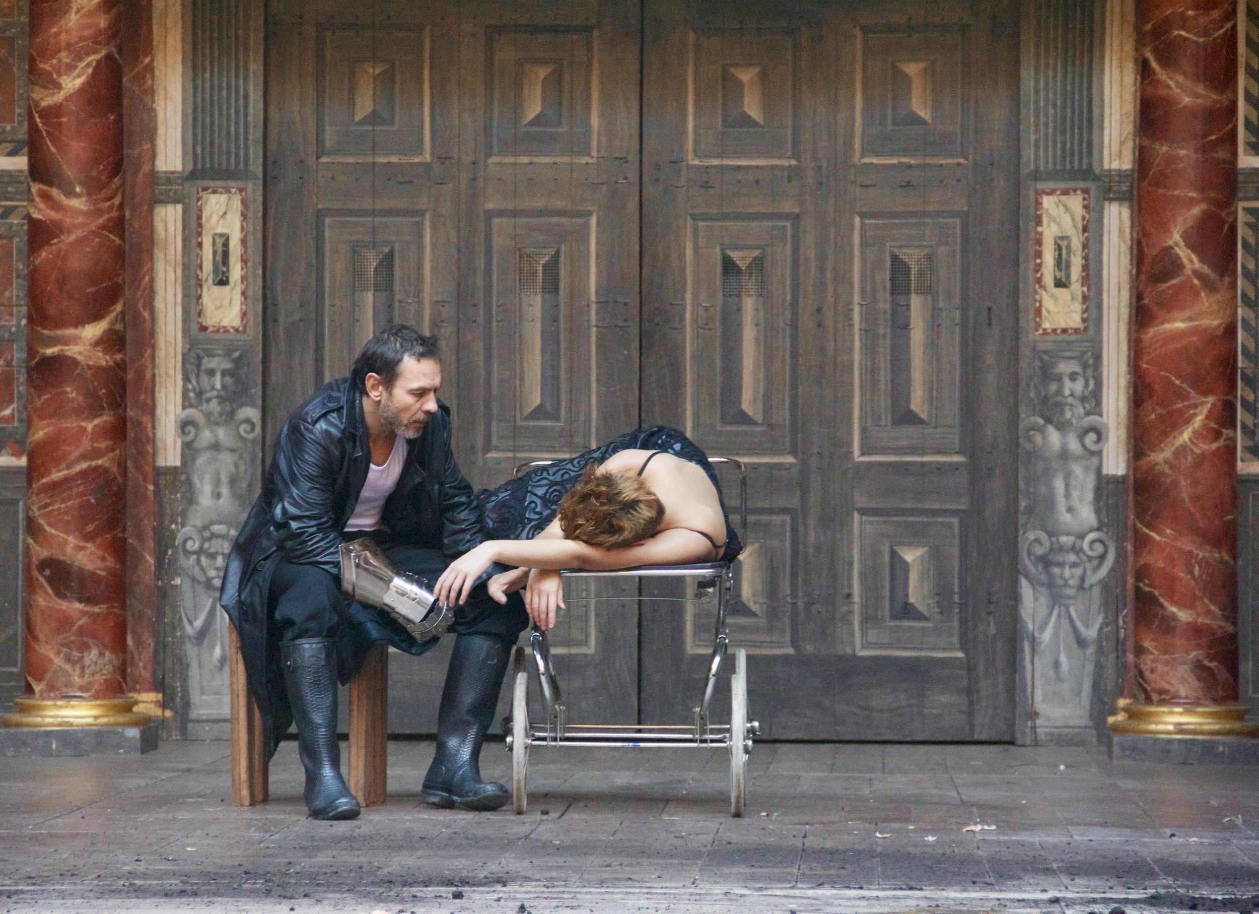 A man with dark hair and a beard wearing a black leather jacket looks down at a woman with curly red hair, dressed in black, lying on a metal stretcher. The woman appears to be unconscious with her head resting on her folded arms. They are positioned in front of a large, ornate wooden door with decorative columns on each side, set in a stone wall with marble-like columns and detailed carvings.