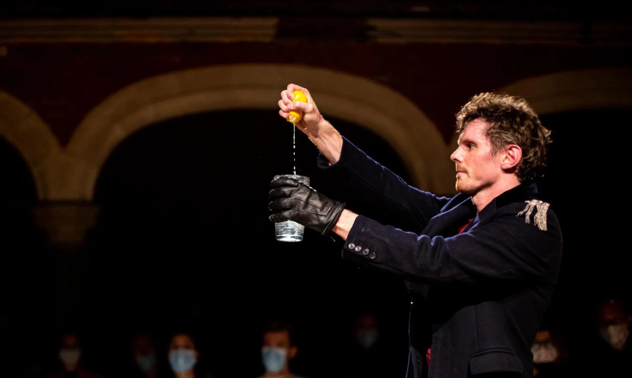 A man with curly hair in a dark jacket and gloves is pouring water from a yellow lemon-shaped container into a glass, with an audience wearing masks visible in the background.