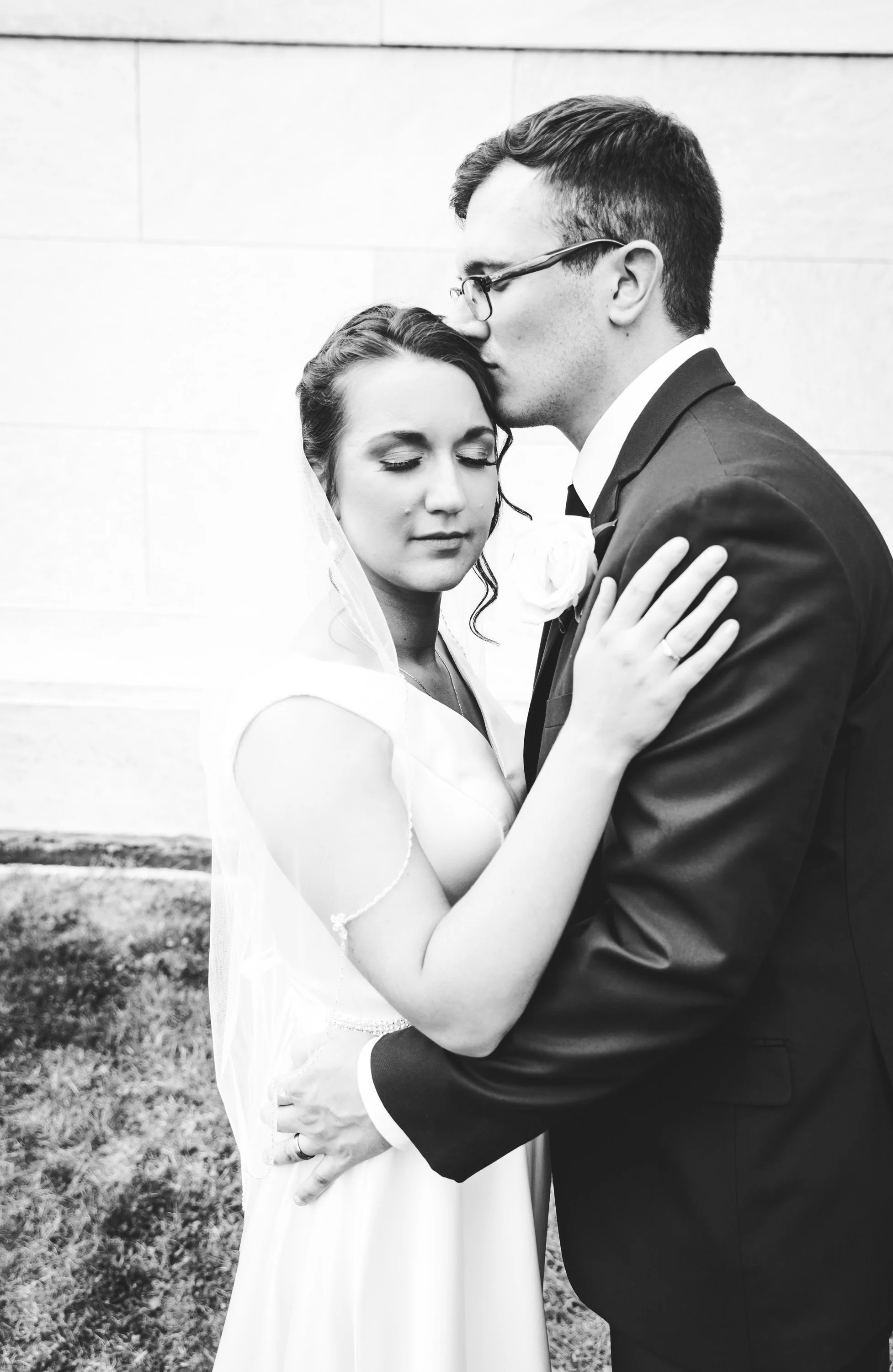Black and white photo of a bride and groom embracing. The groom is kissing the bride on the forehead while she has her eyes closed and her hand resting on his shoulder.