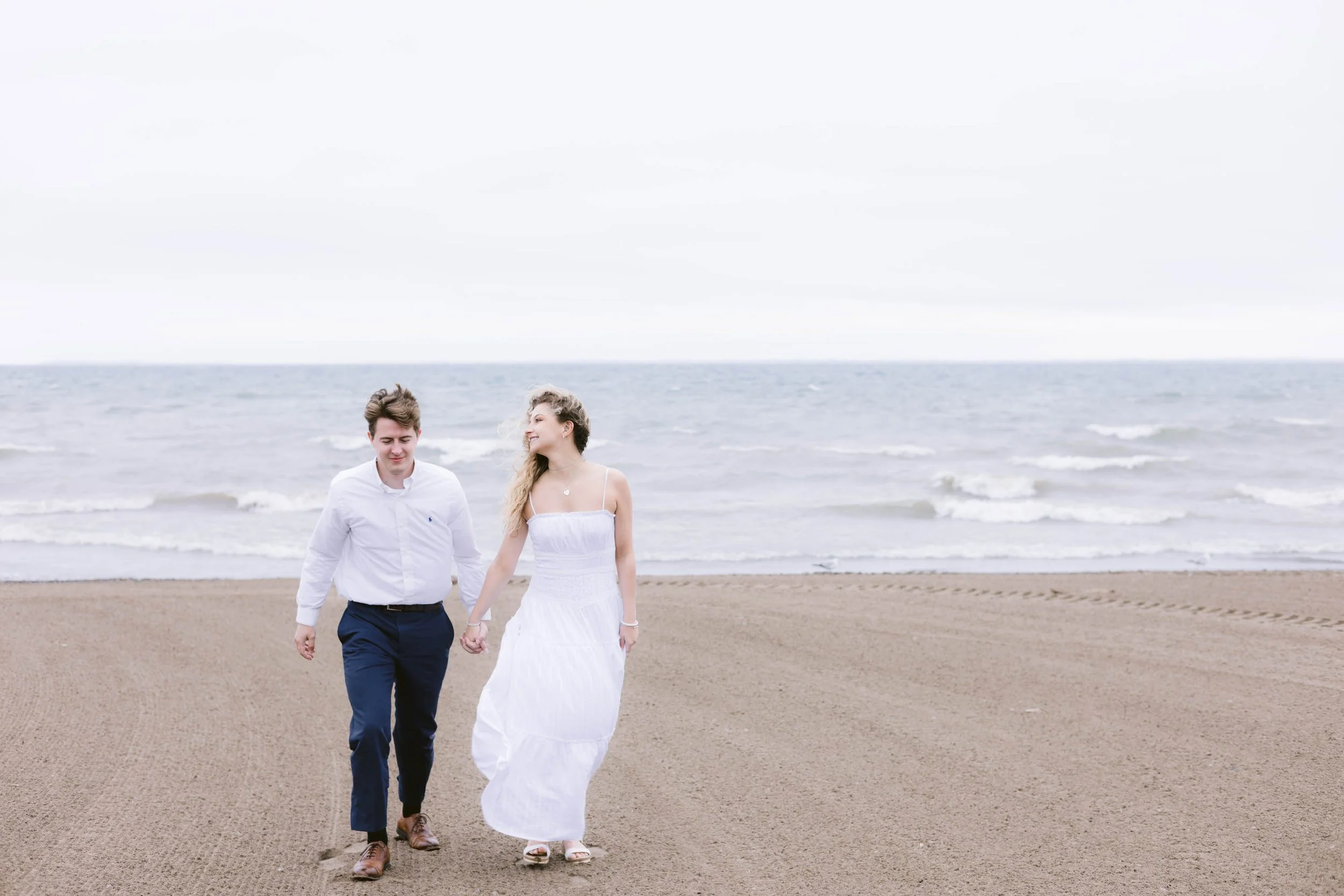 Young couple walking hand in hand on the beach near the ocean, with waves and overcast sky in the background.
