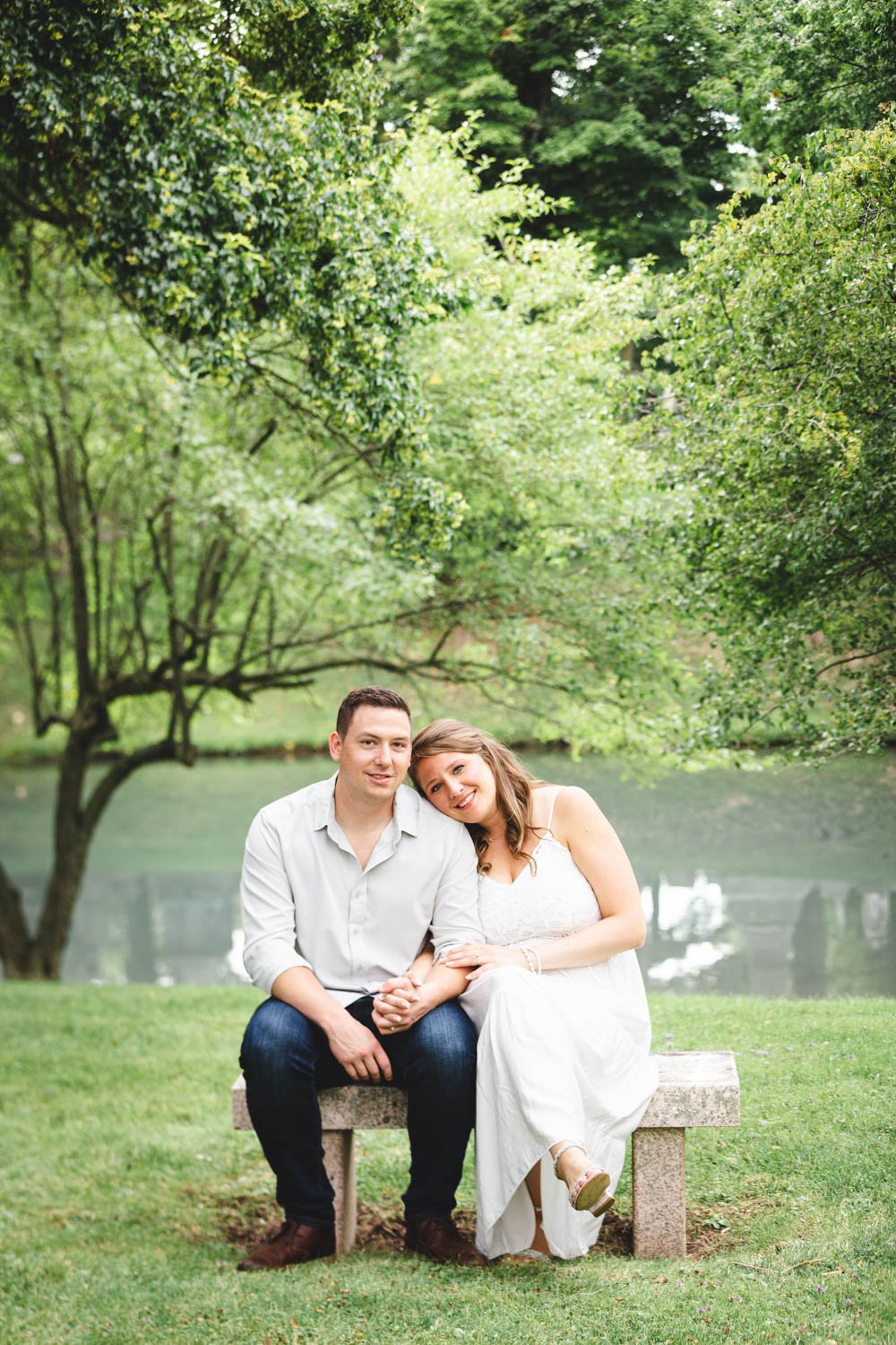 A young couple sitting on a stone bench in a lush, green park near a pond, smiling and leaning against each other.