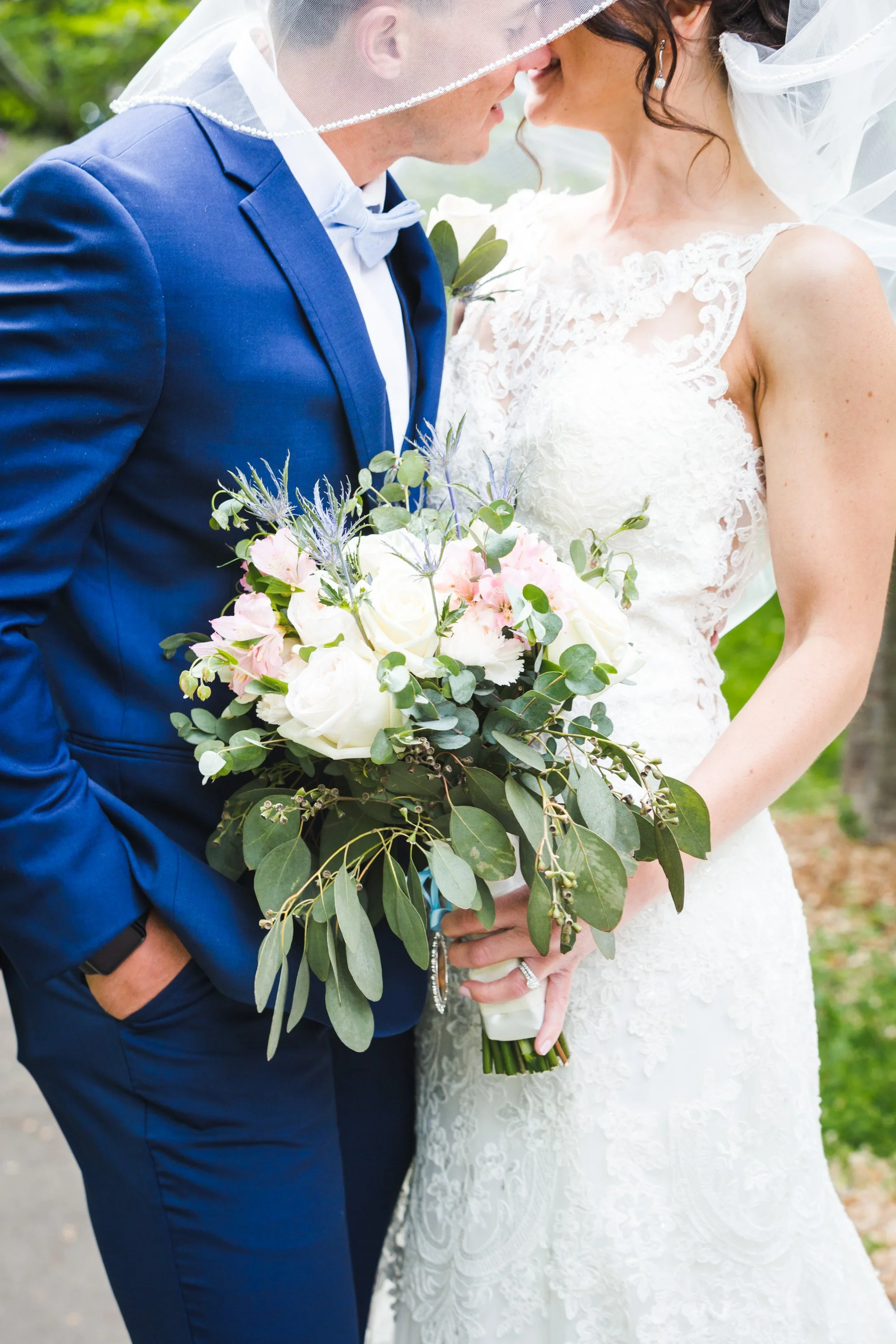 A bride and groom with their faces touching, holding a bouquet of white and pink flowers with greenery, on a green outdoor background.