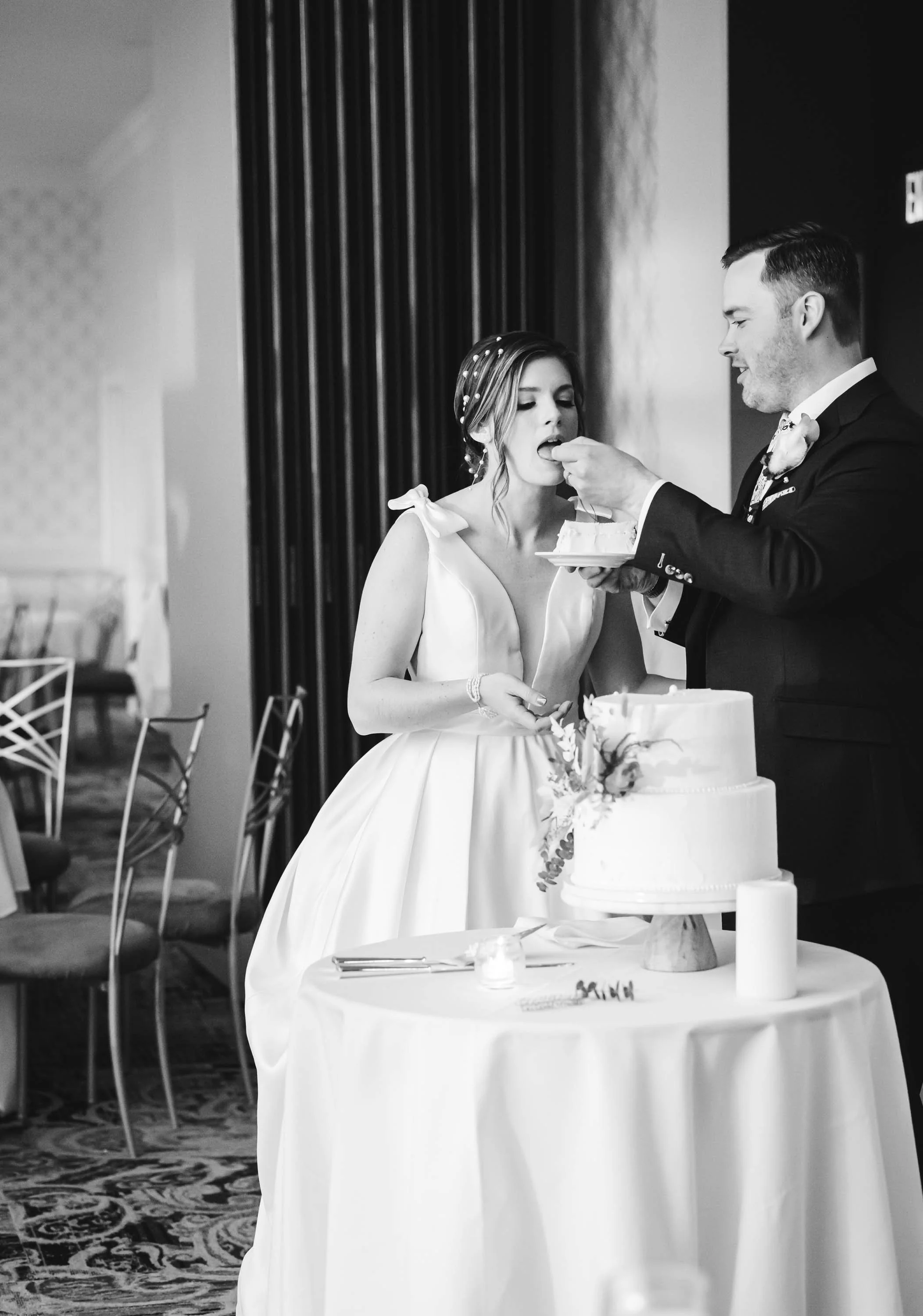A bride and groom at a wedding cake cutting, with the groom feeding the bride a piece of cake.
