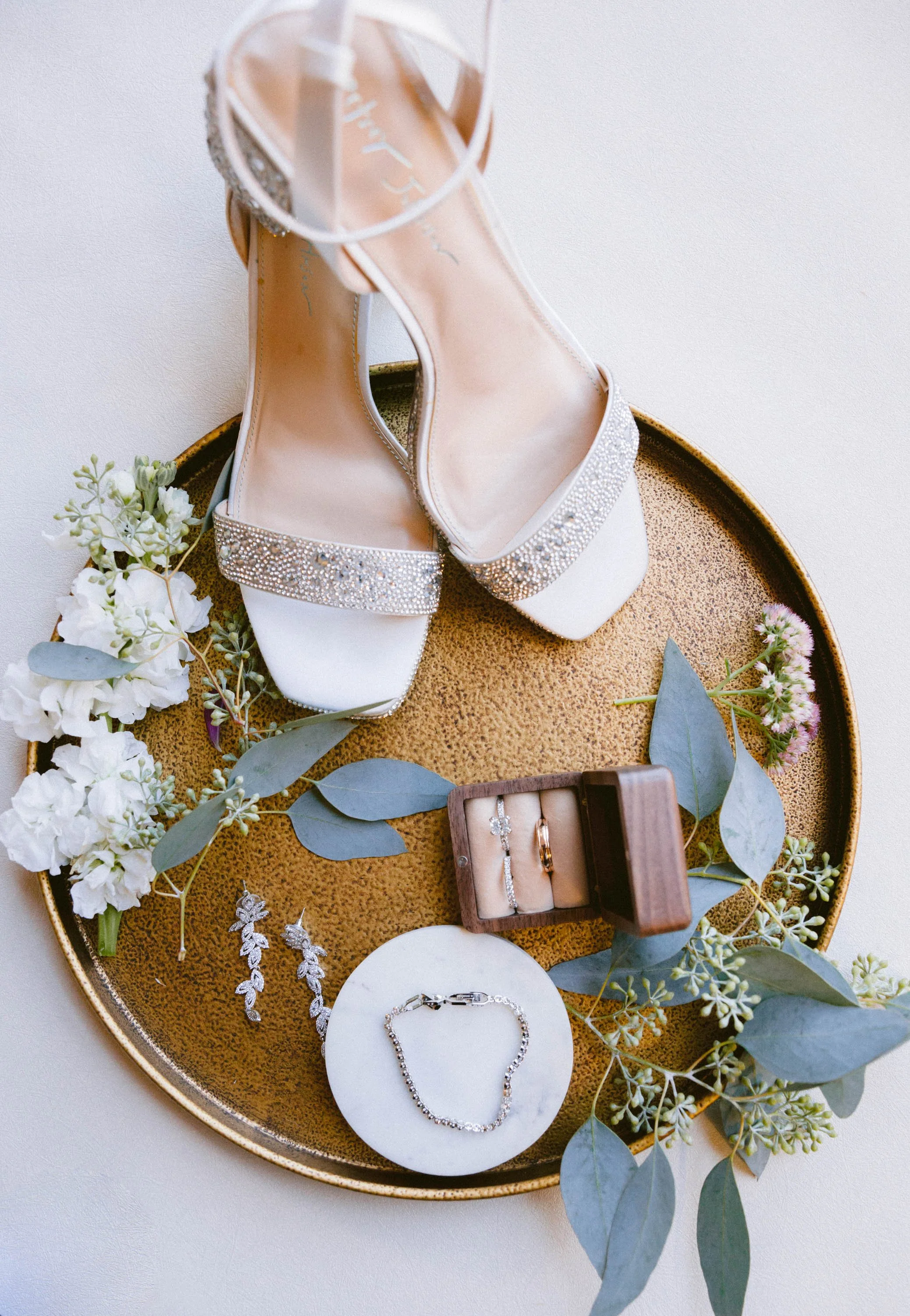 A gold tray holding white high-heeled shoes with rhinestone embellishments, a small wooden box with jewelry including rings and earrings, a bracelet on a white circular dish, surrounded by white and green flowers with eucalyptus leaves.