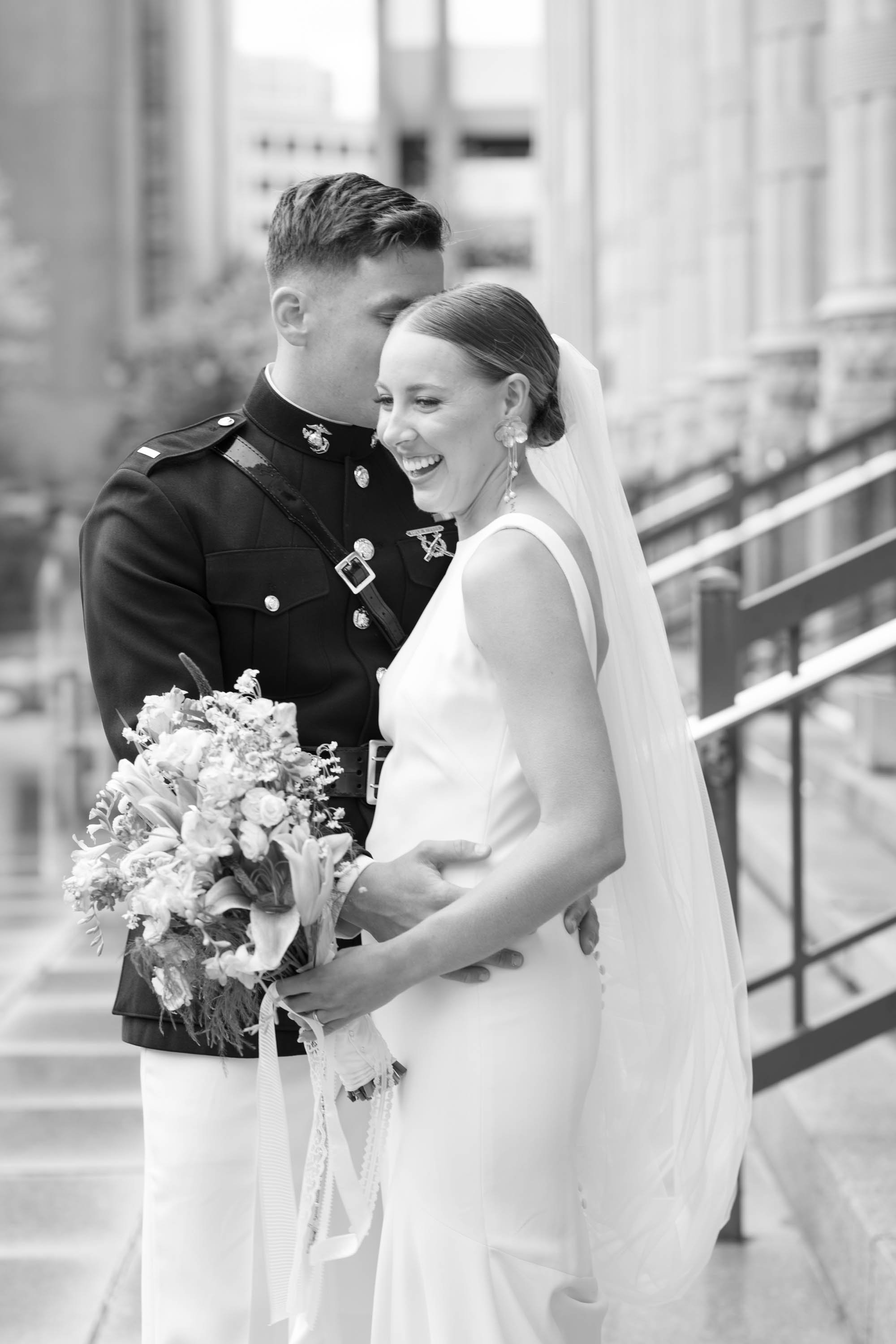 A smiling bride and groom in wedding attire sharing a moment outdoors in black and white.