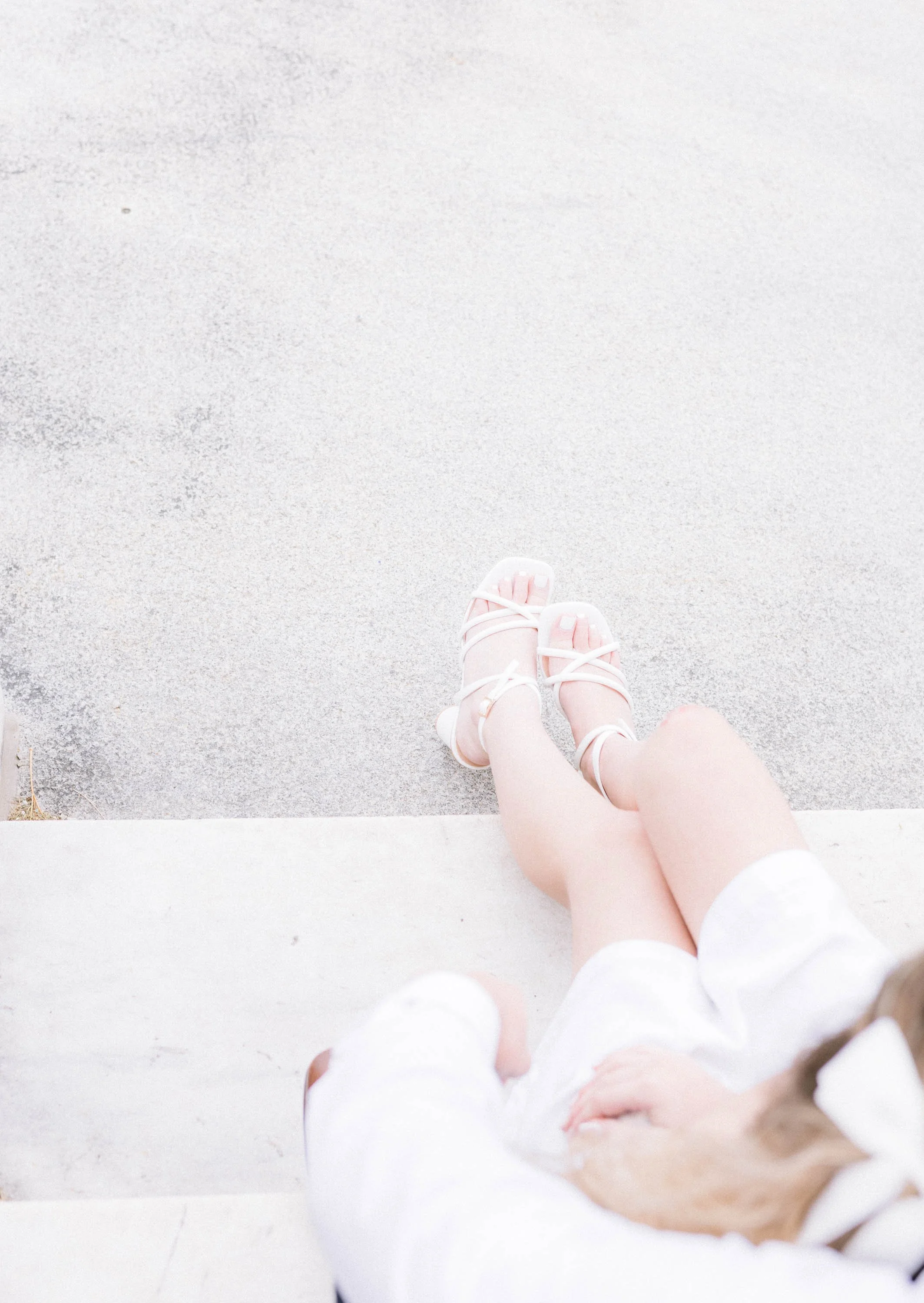 Close-up of a person's legs and feet sitting on a white step, wearing pink sandals and white tights, with a concrete ground in the background.