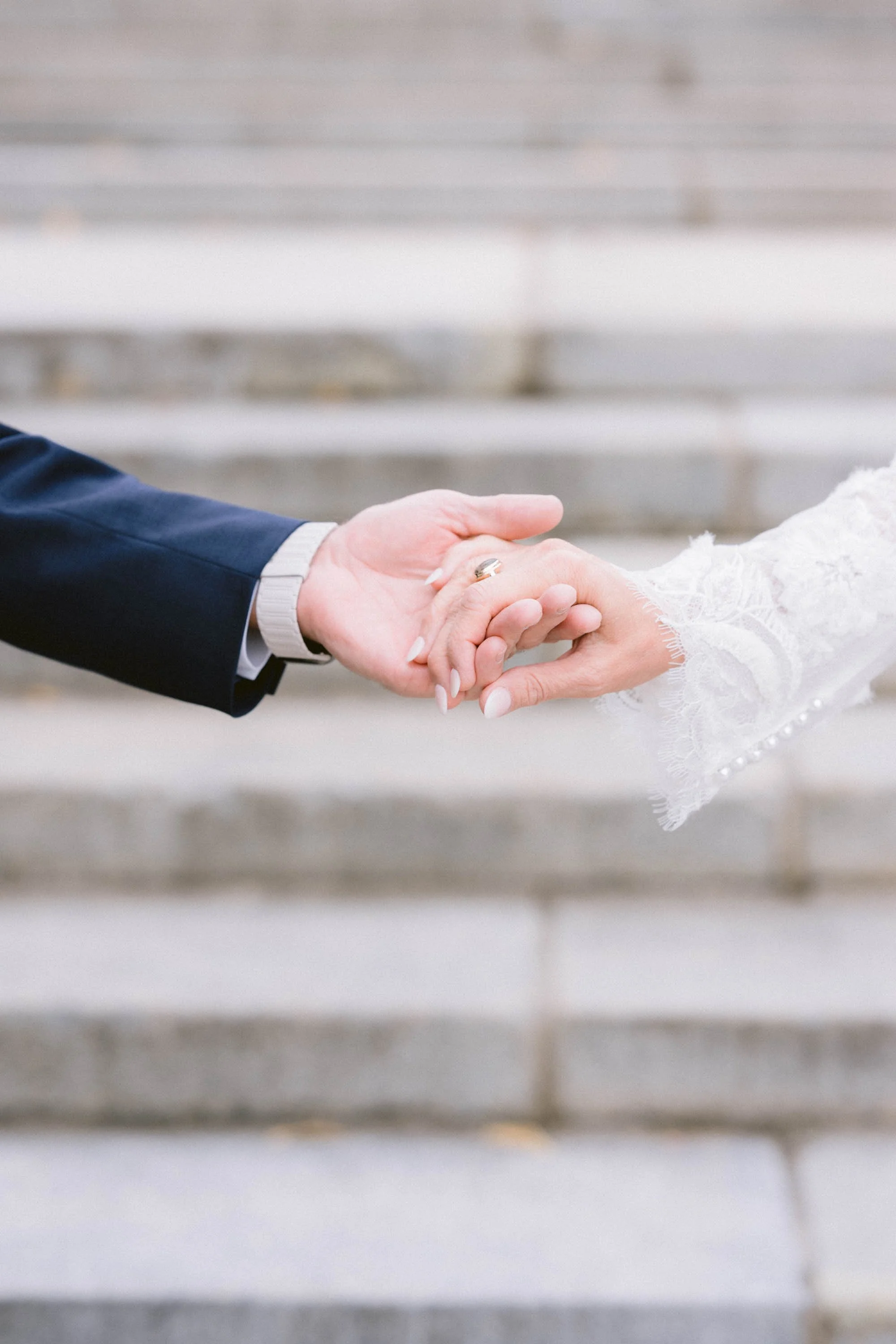 Close-up of a bride and groom holding hands, with the groom wearing a dark suit and the bride in a lace dress, on stone steps.